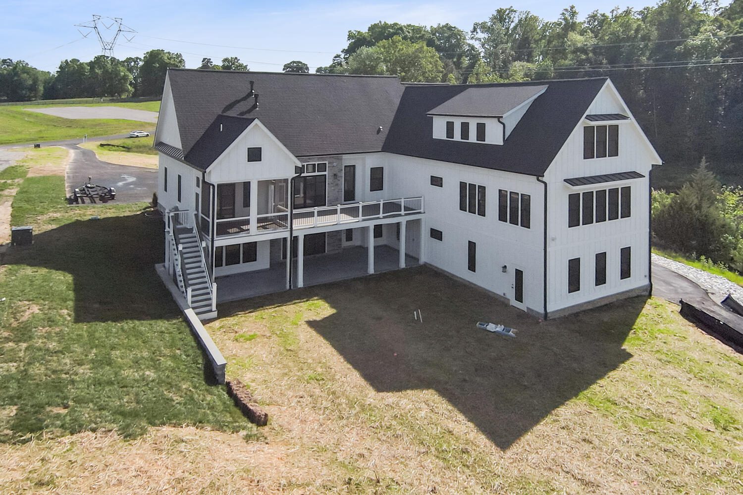 A large two-story white house with a dark roof, surrounded by grass and trees, features multiple windows and a spacious elevated deck.