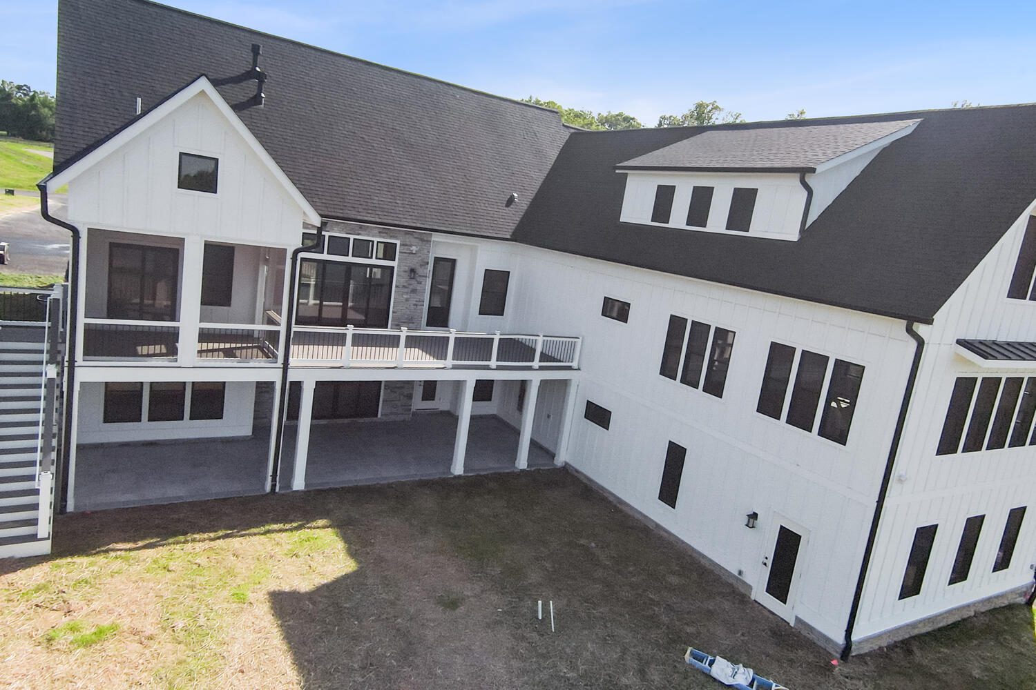 A large white house with a dark roof features multiple windows, a screened porch, and a deck. The backyard is partially shaded and grassy.