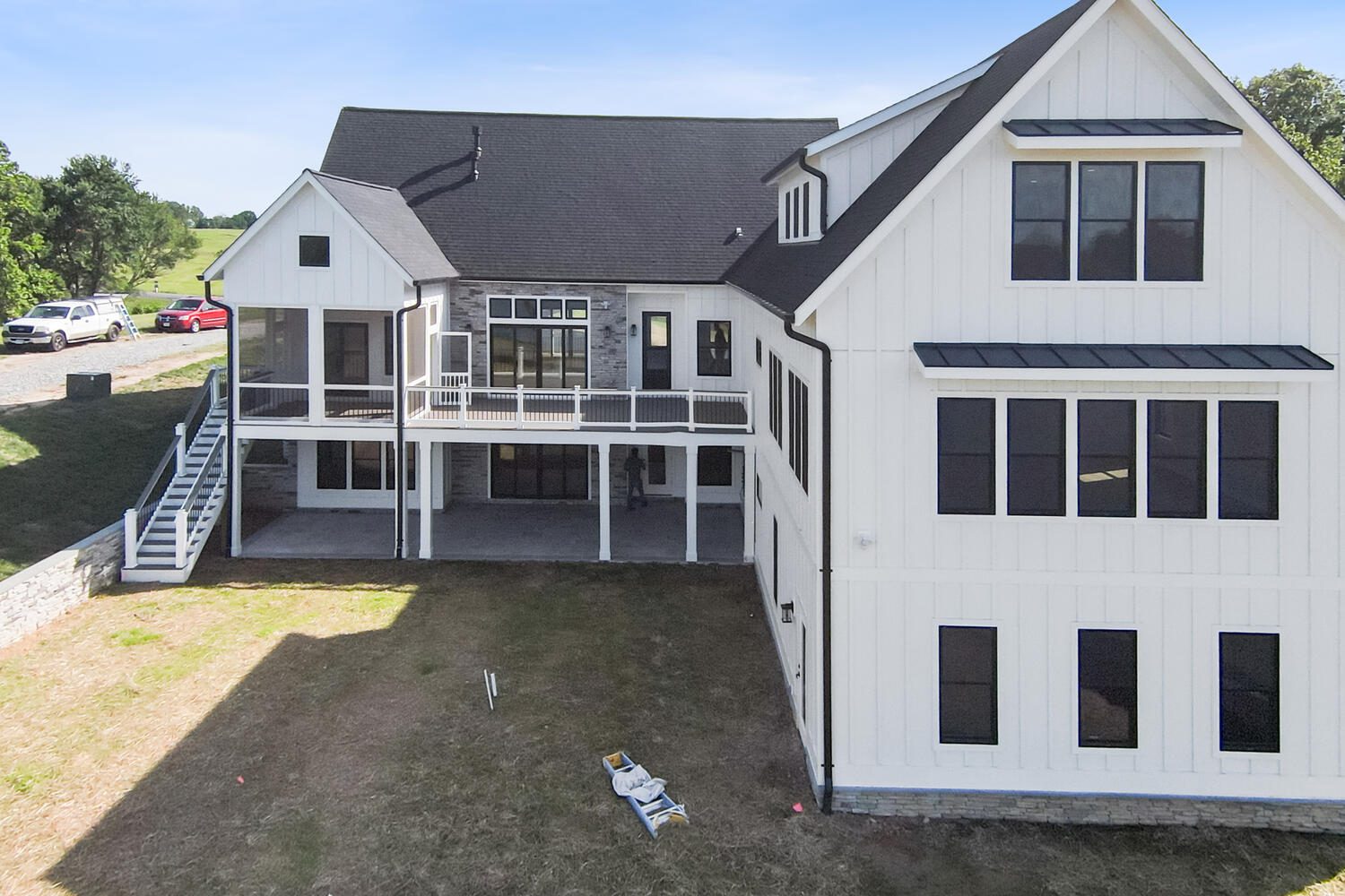 Large white house with black roof, multiple windows, and wraparound porch. Grassy yard in the foreground, with several vehicles parked on the driveway to the left.
