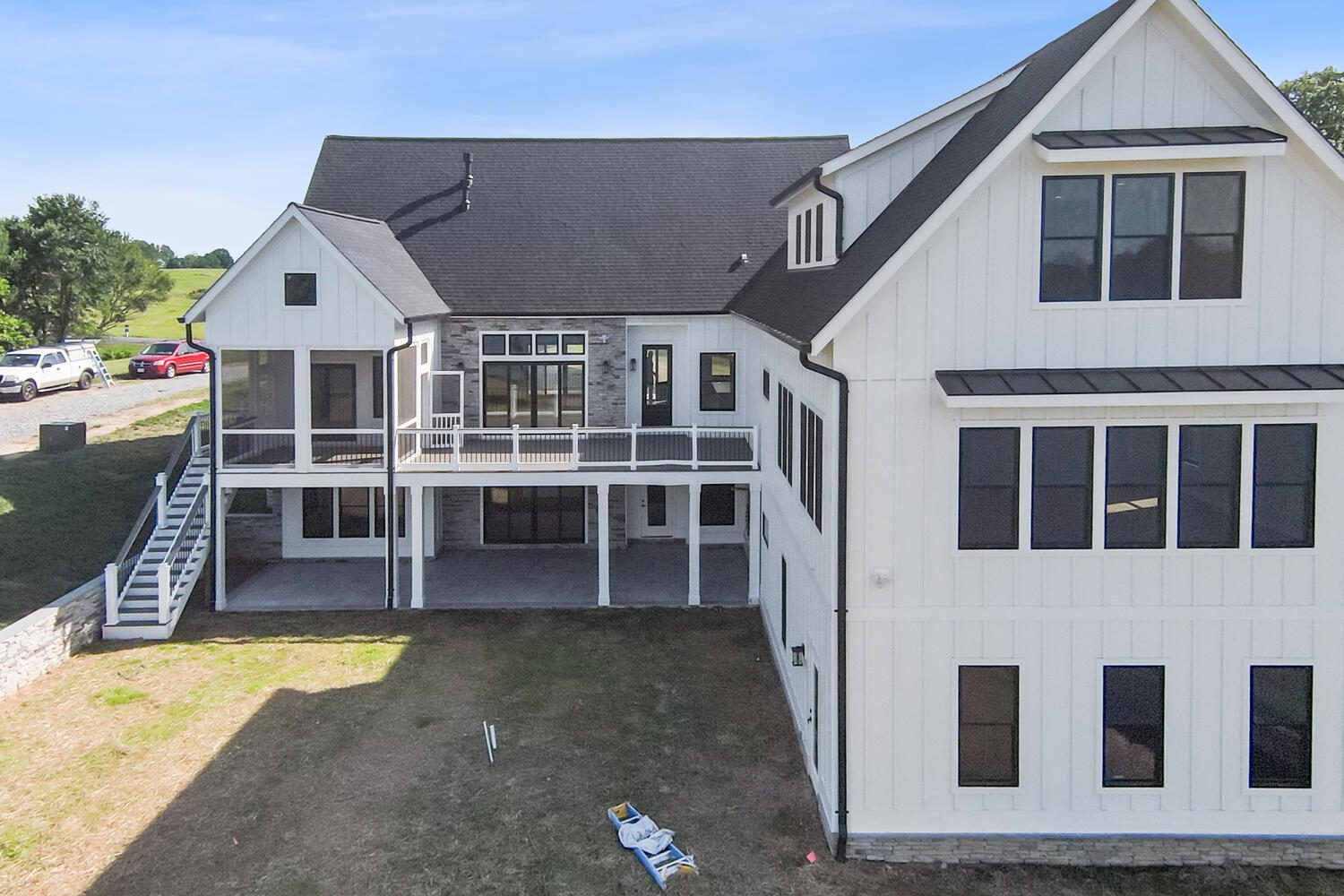 A large white house with multiple windows, black roof, and a covered porch. A grassy area and stairs lead to the upper balcony. Several vehicles are parked on the side.