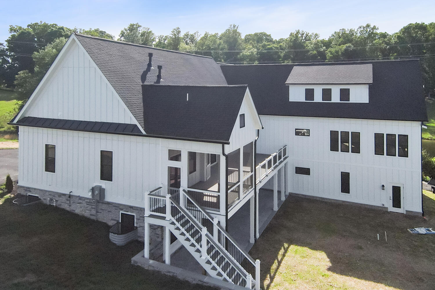 Two-story modern farmhouse with white siding, black roof, and multiple windows. Features an outdoor staircase leading to a covered porch, set against a backdrop of trees and grass.