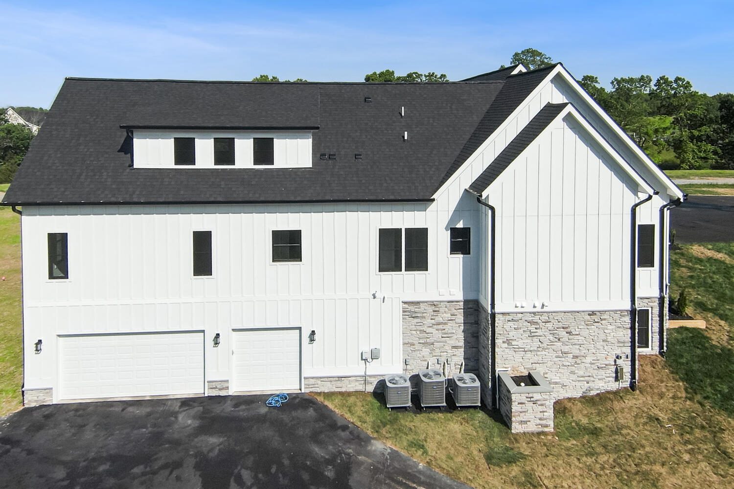A modern two-story house with a white exterior, black roof, three garages, stone accents, and visible air conditioning units on the side.