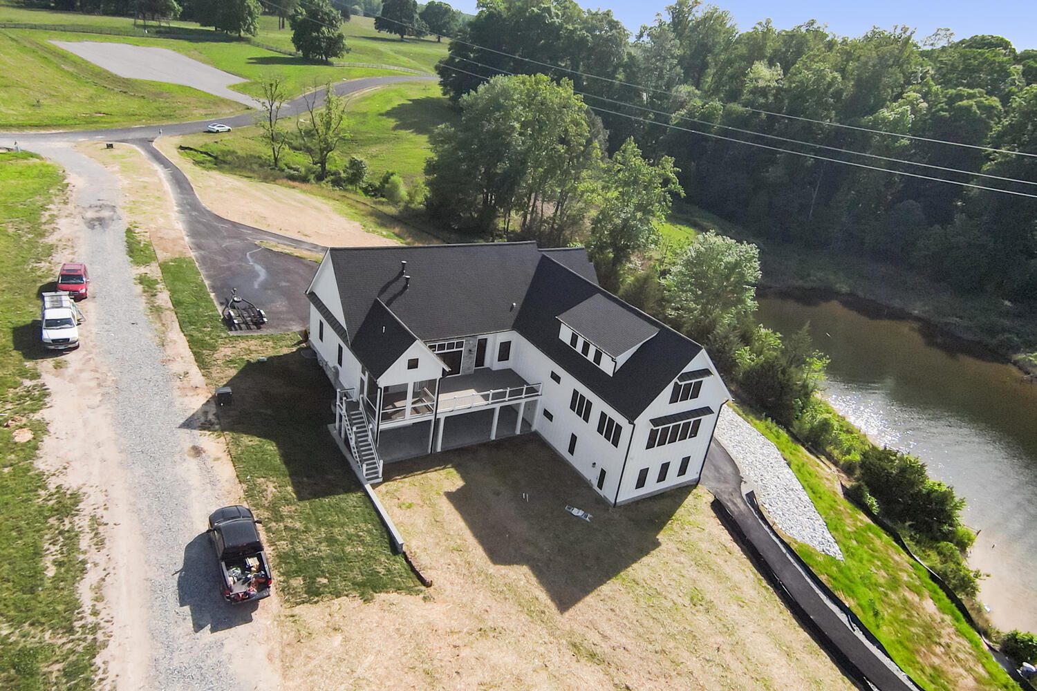 Aerial view of a large, white modern house with a black roof, located near a pond. Several vehicles are parked on the surrounding gravel driveway and road.