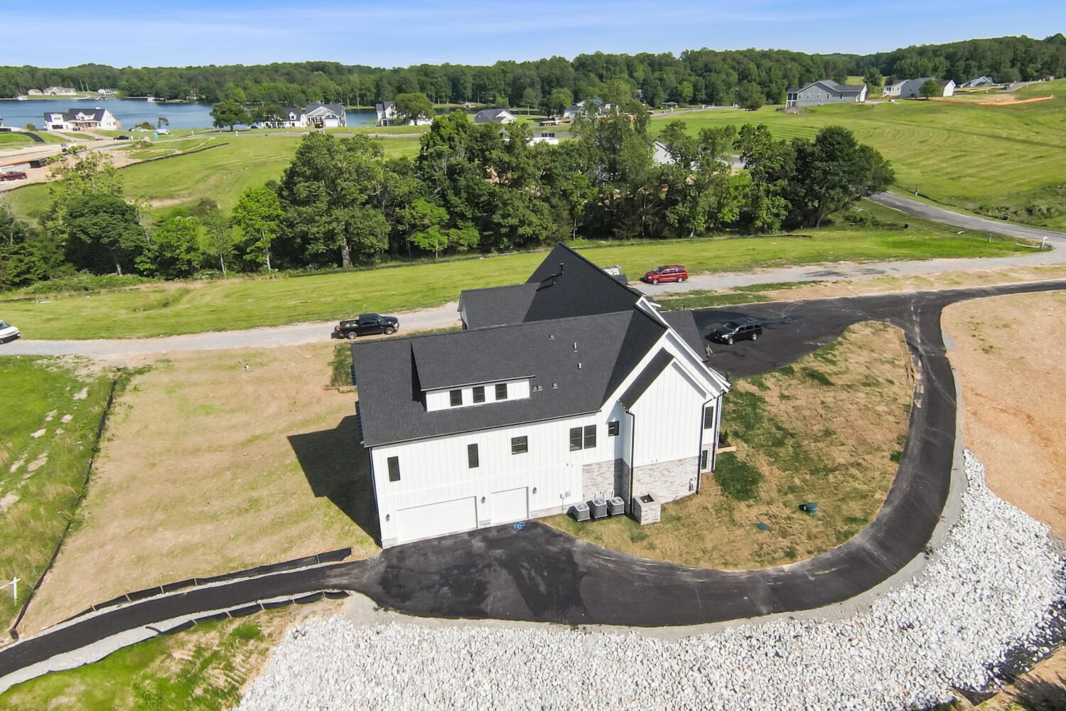 Aerial view of a white house with a dark roof, surrounded by grassy fields and a paved driveway. A lake and scattered homes are visible in the background.