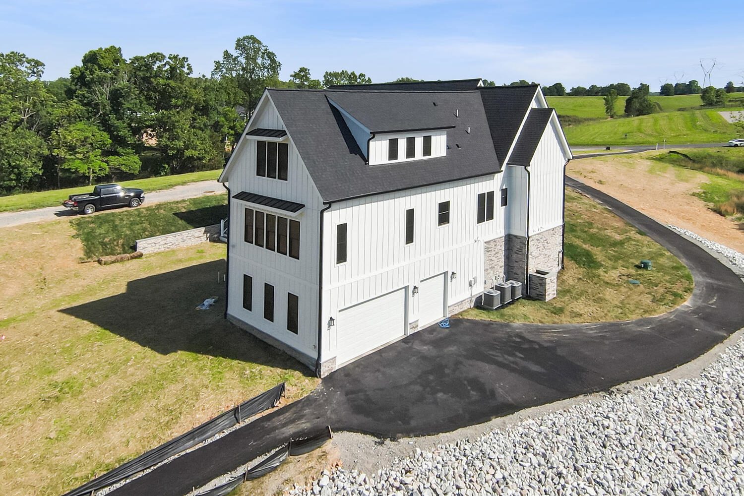 Large modern farmhouse with black roof, white siding, and stone accents, situated on a hill with a curved driveway, surrounded by grass and trees.