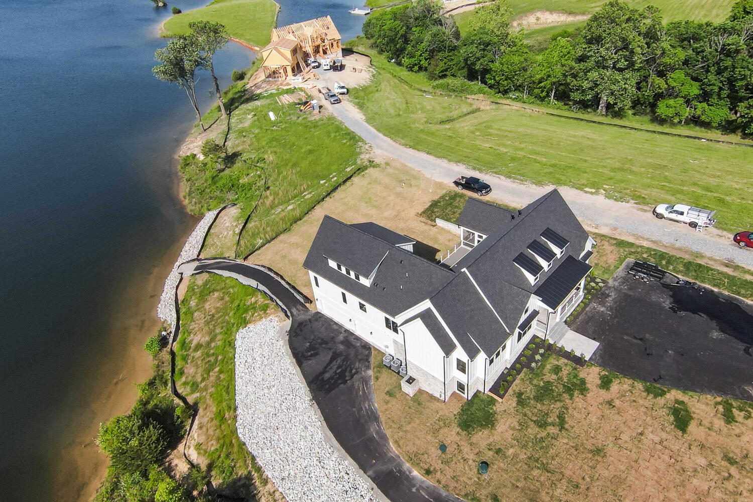 Aerial view of a large house by a lake, with a driveway, nearby trees, and another building under construction.