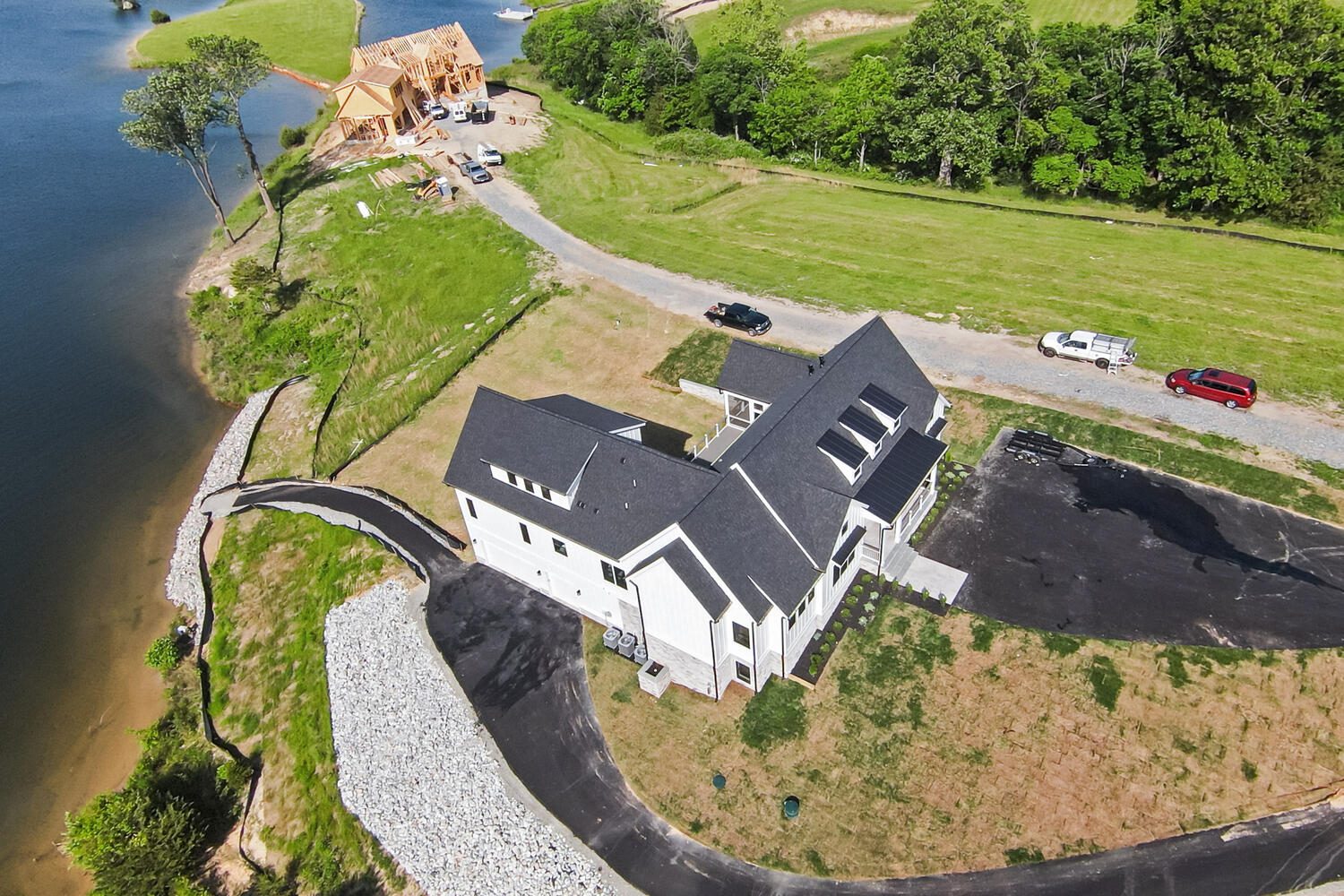 Aerial view of a modern white house with dark roof near a lake, surrounded by greenery. A winding road leads to the house. A wooden structure is under construction nearby. Several vehicles are visible.