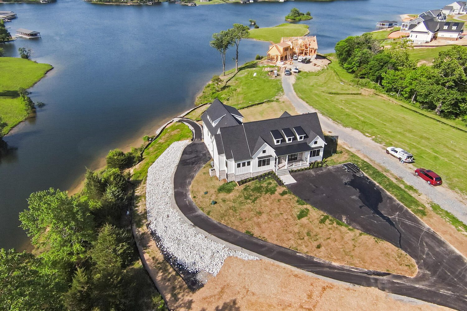 Aerial view of a house with a dark roof by a lakeside, partially surrounded by a white rock embankment. A red car is parked on a nearby driveway. Construction is visible in the background.