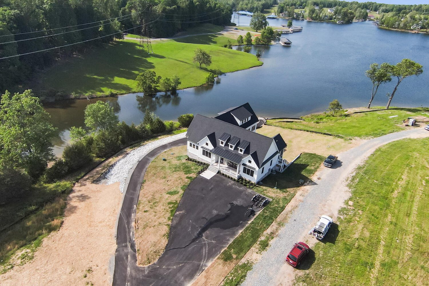 Aerial view of a large house near a lake, surrounded by trees. A paved driveway leads to the property, with several vehicles parked nearby.