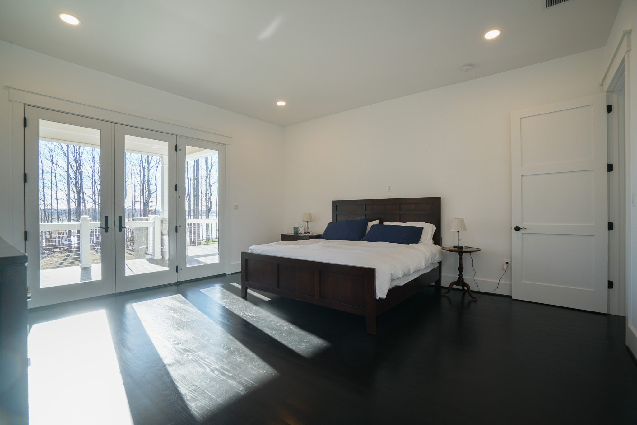Bedroom with a wooden bed, white bedding, and two bedside tables. Sunlight streams in through glass doors, illuminating the dark wood floor. White walls and recessed lighting create a minimalist feel.