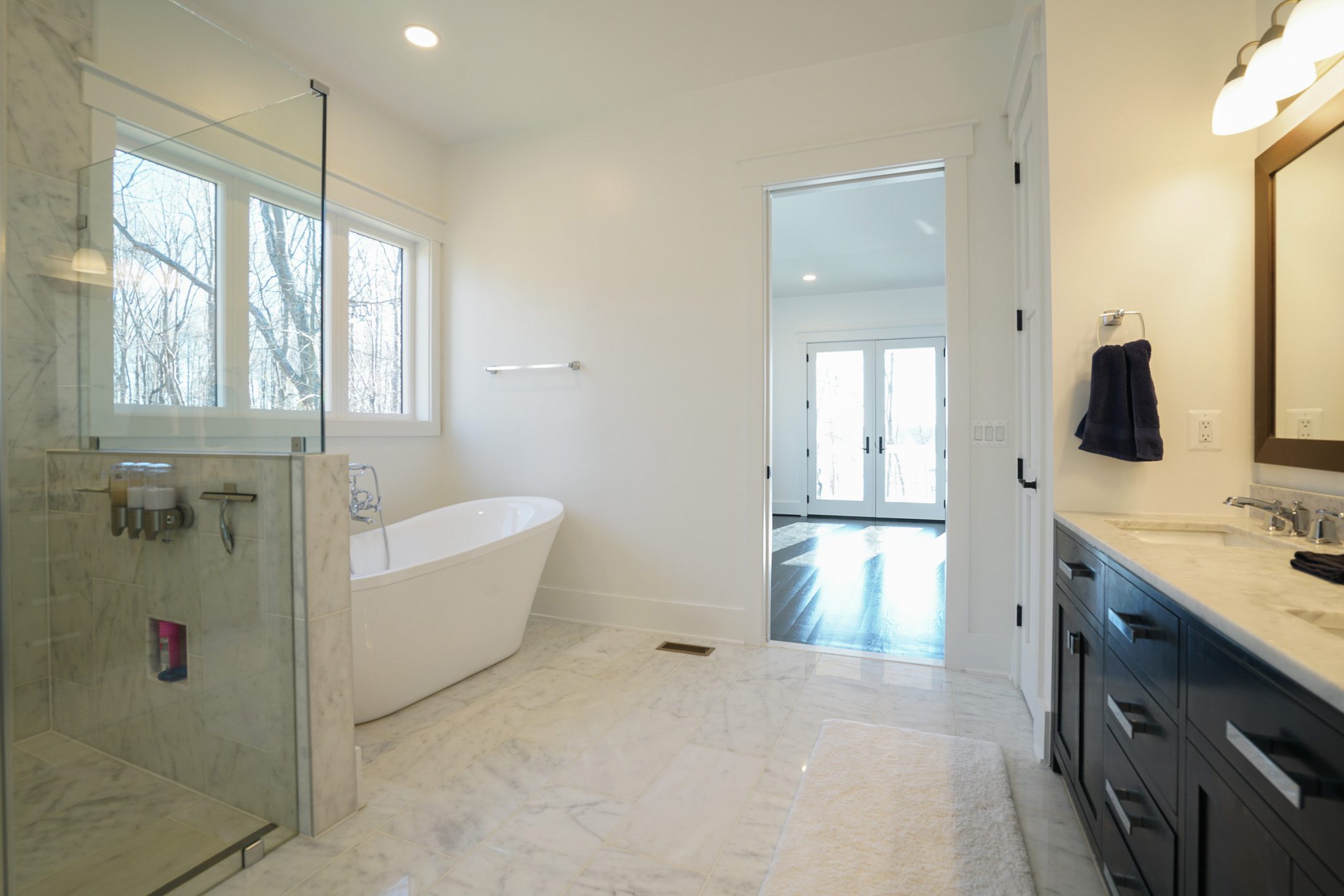 Modern bathroom with a freestanding bathtub, glass-enclosed shower, and dark wood vanity. Large windows provide natural light. An open doorway leads to a room with wooden flooring.