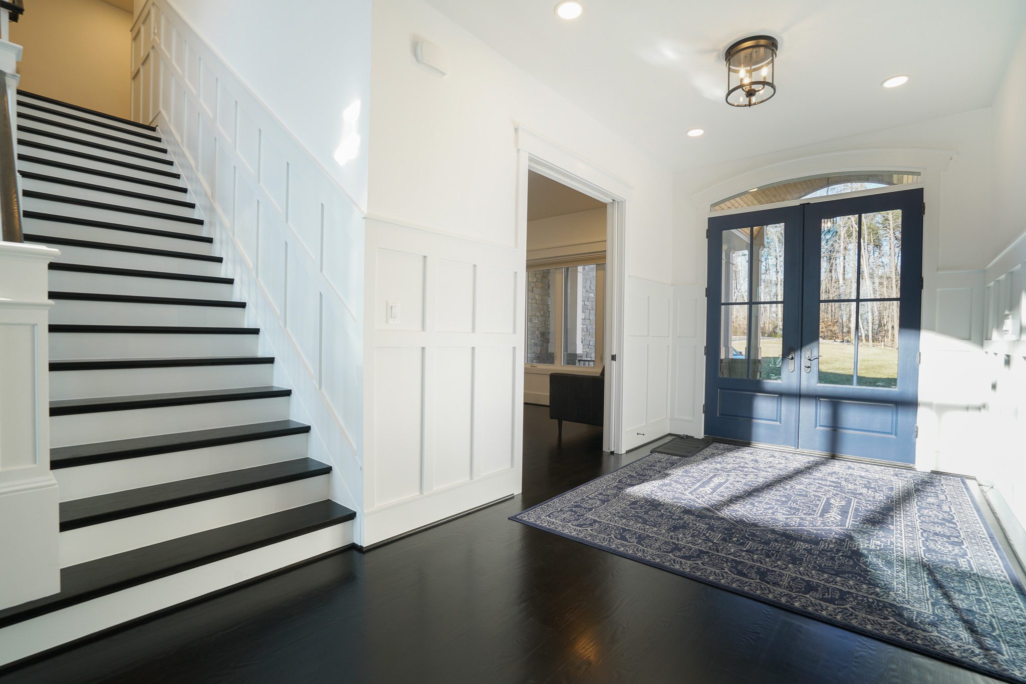 Bright foyer with large double doors, dark wood floor, and white walls. Staircase on the left with white risers and dark treads. A blue patterned rug lies on the floor.