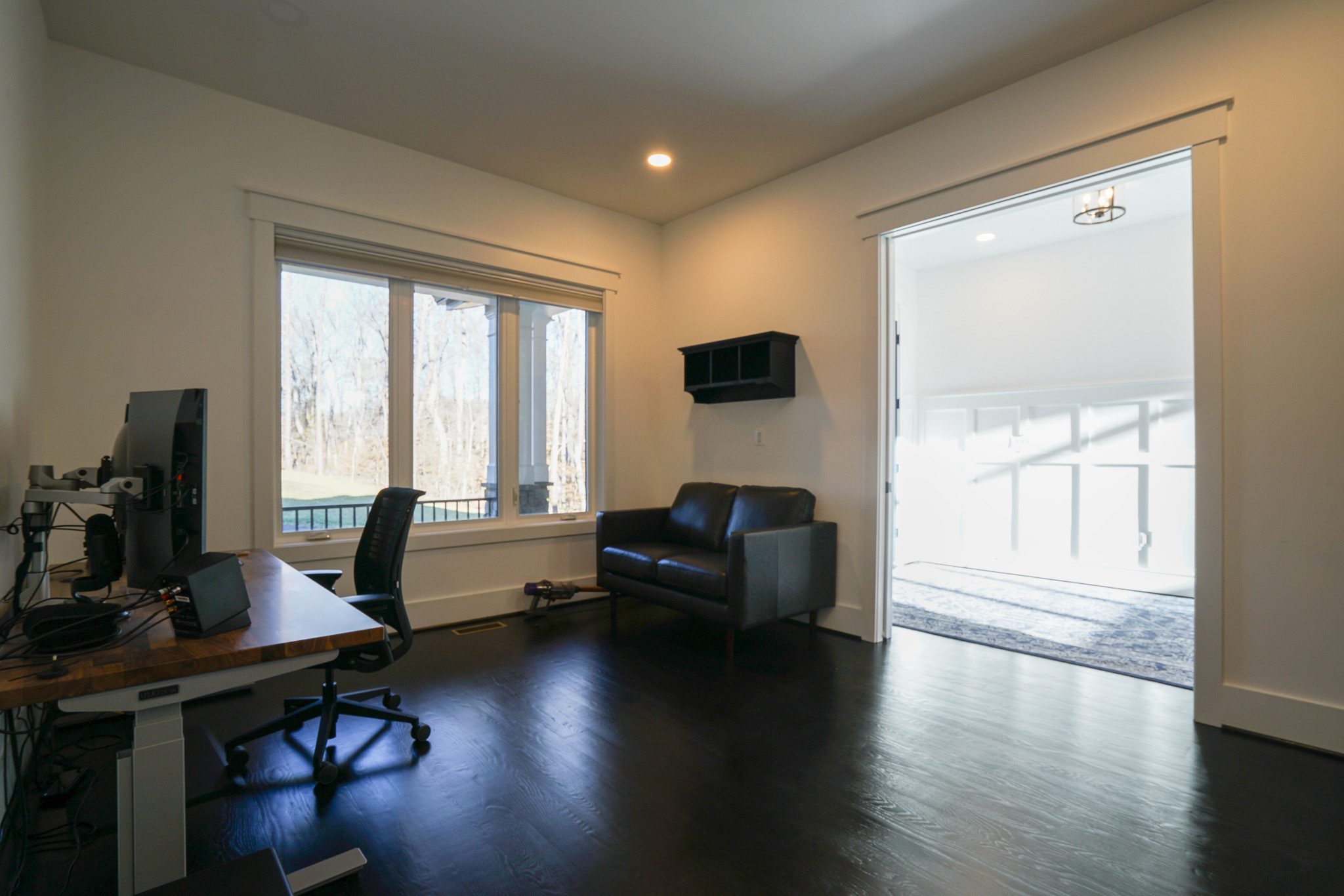 A home office with a desk, chair, and computer on the left. A black couch is in the center. Large window on the left wall. Sliding doors open to a bright adjacent room with white walls.