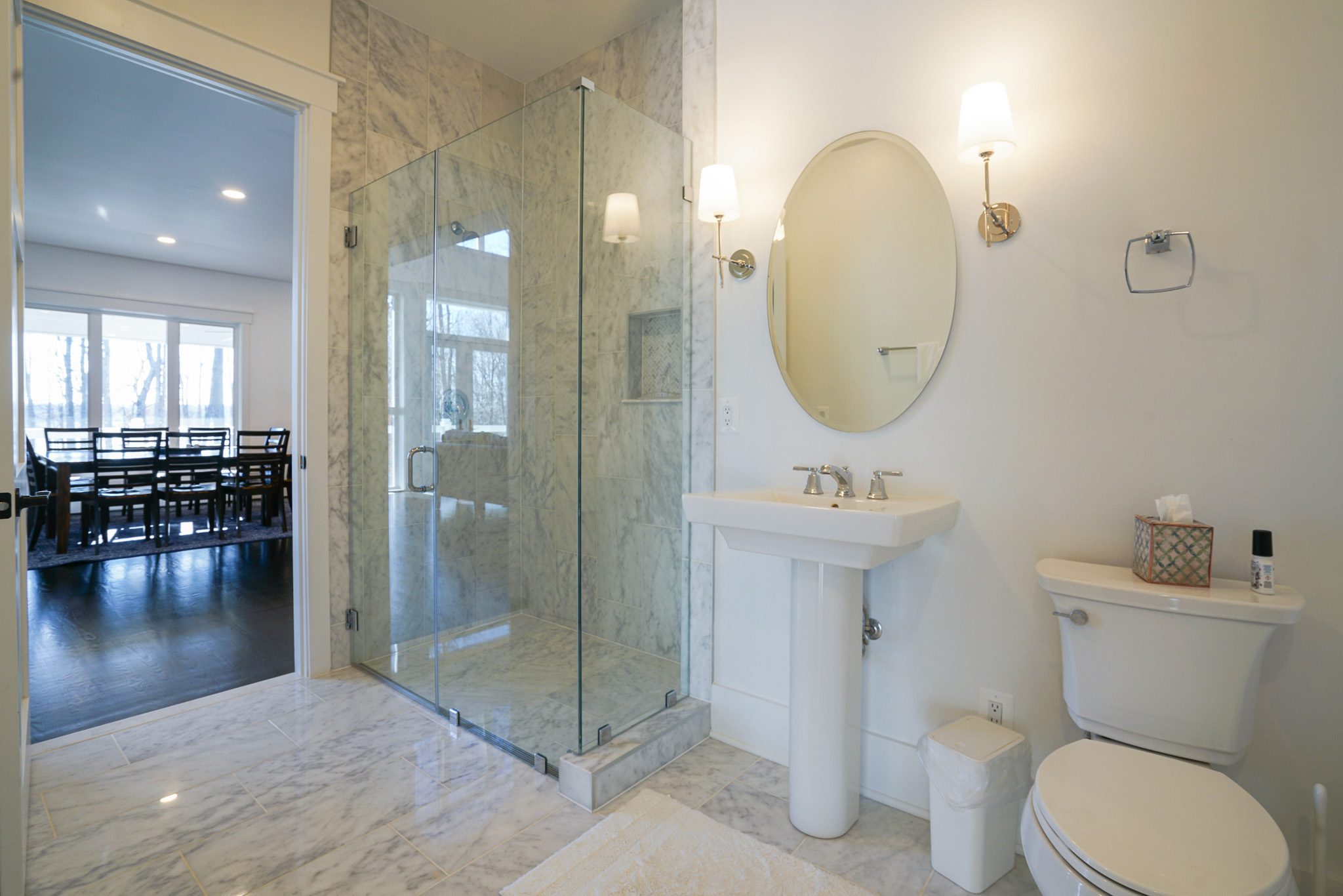 Bathroom with a glass-enclosed shower, oval mirror, pedestal sink, and toilet. Marble tiles on floor and walls. View through door shows a dining room with a table and chairs.