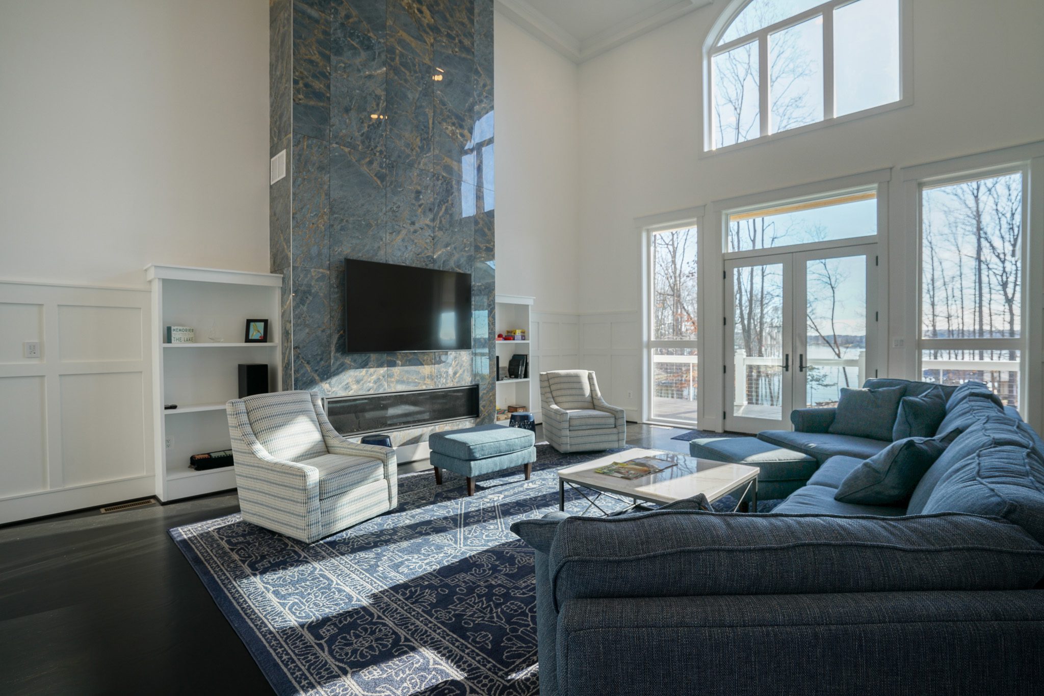 Modern living room with high ceilings, large marble fireplace, wall-mounted TV, blue sectional sofa, two striped armchairs, a blue ottoman, and a rug. Large windows provide natural light.
