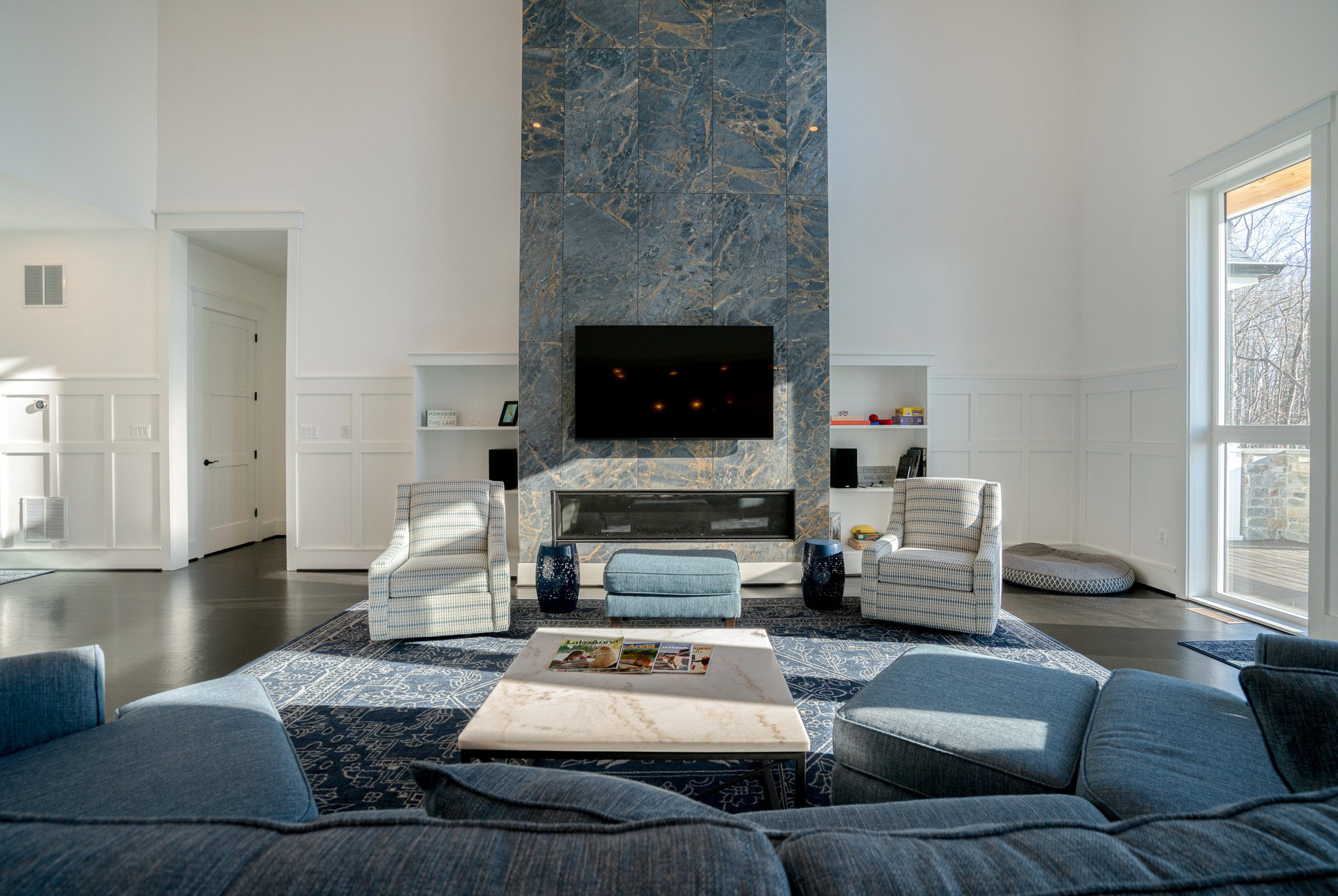 Spacious living room with blue sofas, armchairs, and a central square coffee table. A large TV hangs above a fireplace set in a tall blue marble wall, flanked by white bookcases.