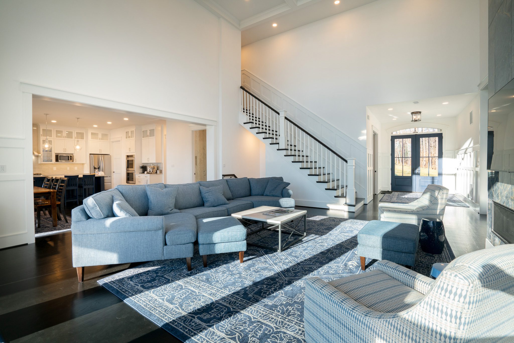 Spacious living room with blue sofas, patterned rugs, and a view into the kitchen. A staircase leads to the upper floor. Sunlight streams in through large windows.