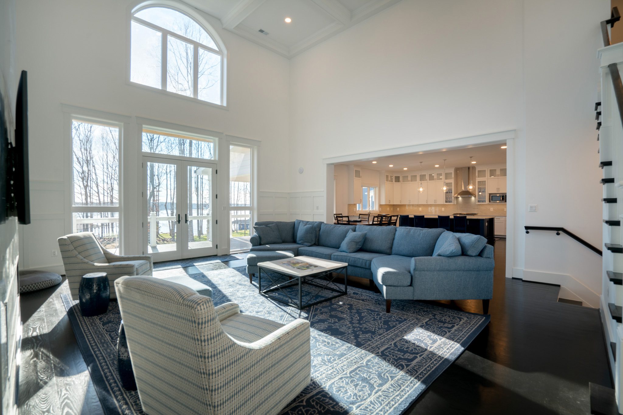 Spacious living room with large windows, a blue sectional sofa, patterned rug, and armchairs. Open layout leads to a bright kitchen with white cabinetry and bar seating.
