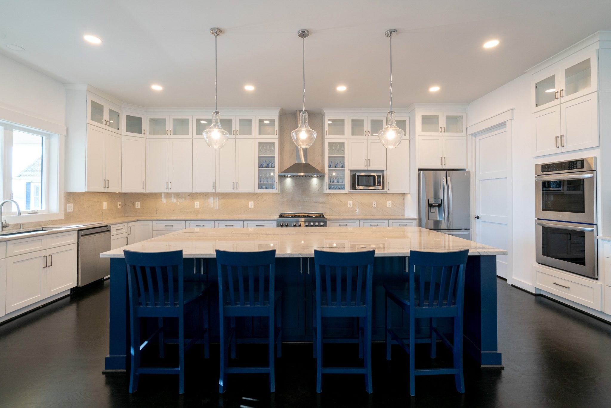 Modern kitchen with white cabinetry, stainless steel appliances, and a large island with dark blue chairs. Three pendant lights hang above the island.