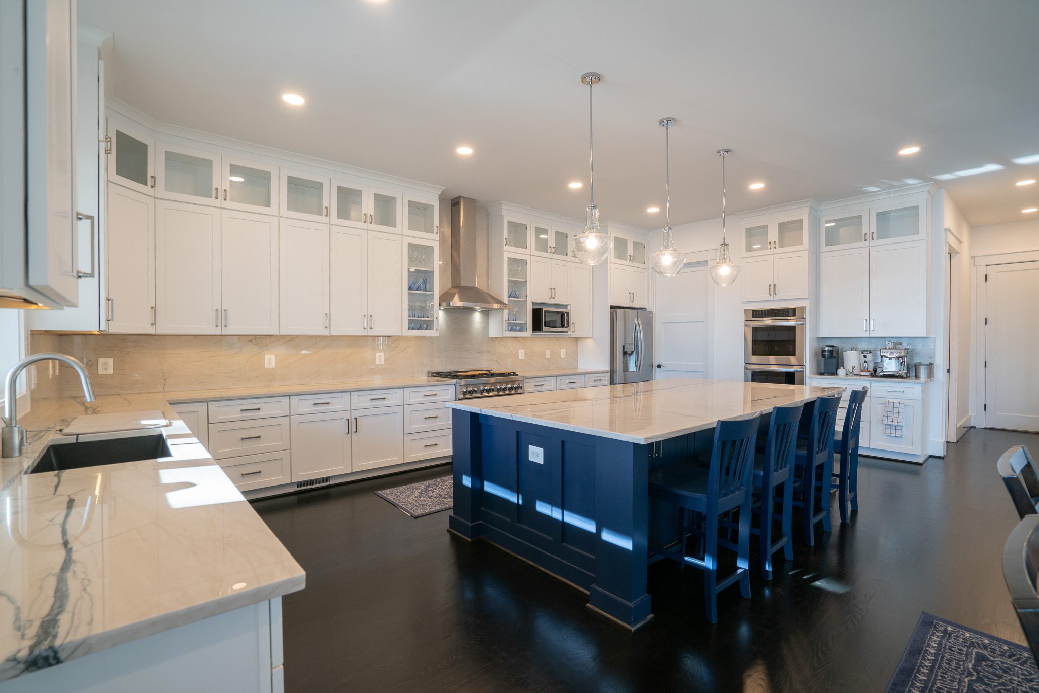 Spacious kitchen with white cabinets, blue island, marble countertops, and three pendant lights. Features stainless steel appliances and a dark hardwood floor.
