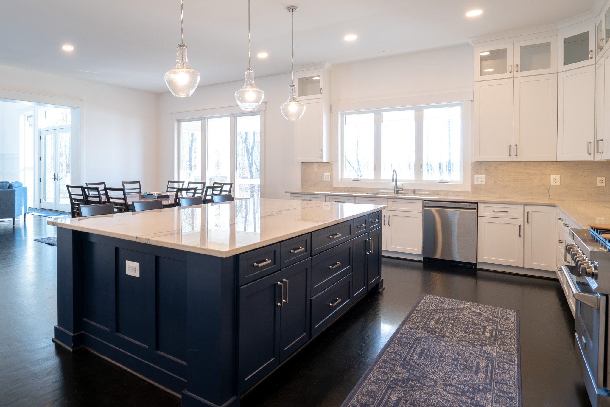 Modern kitchen with white cabinets, dark blue island, stainless steel appliances, and three pendant lights. Dining area with table and chairs is visible in the background.