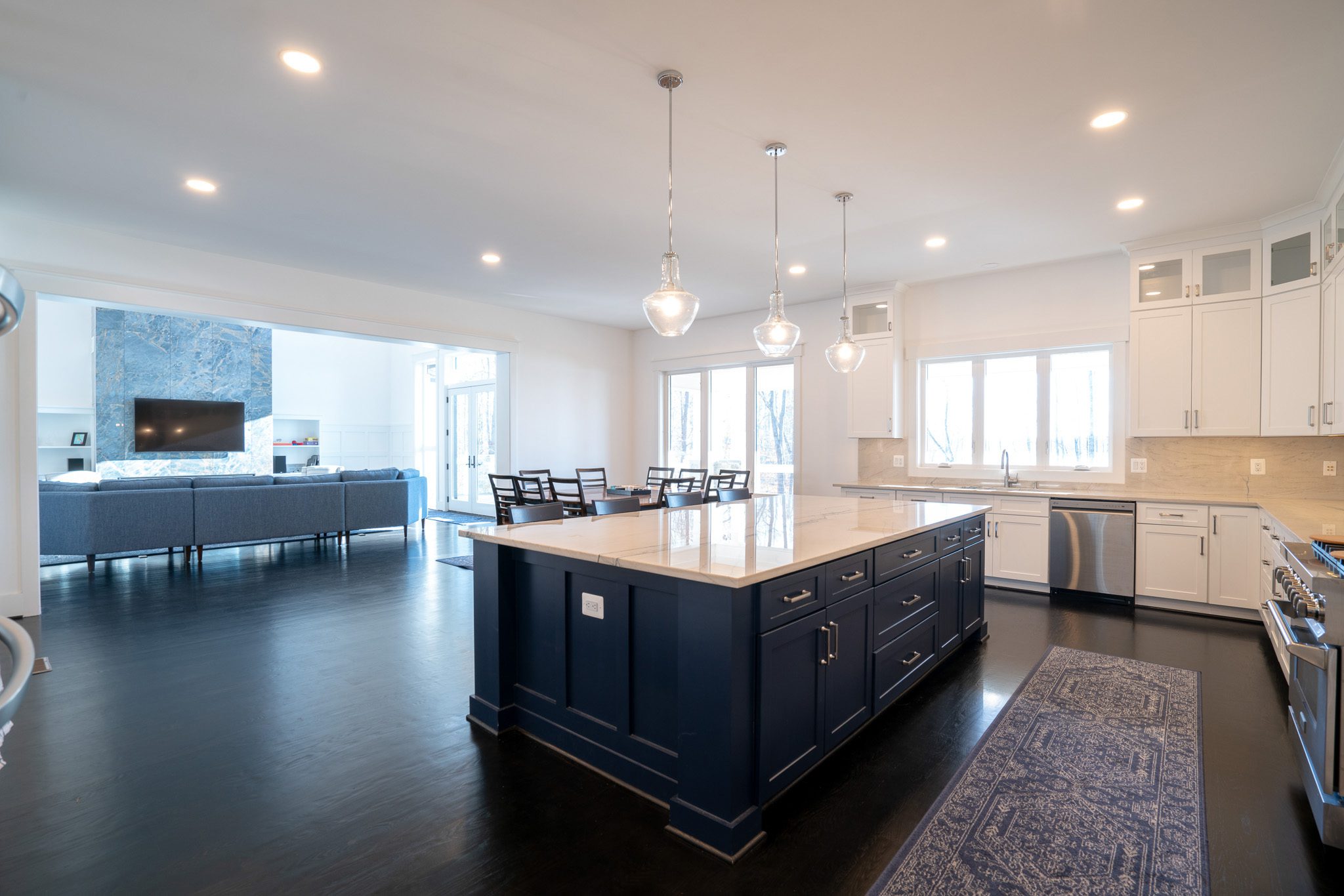 A modern kitchen with a large blue island, marble countertops, pendant lights, and adjacent living area. Dark hardwood floors, white cabinetry, and a visible dining table enhance the open layout.