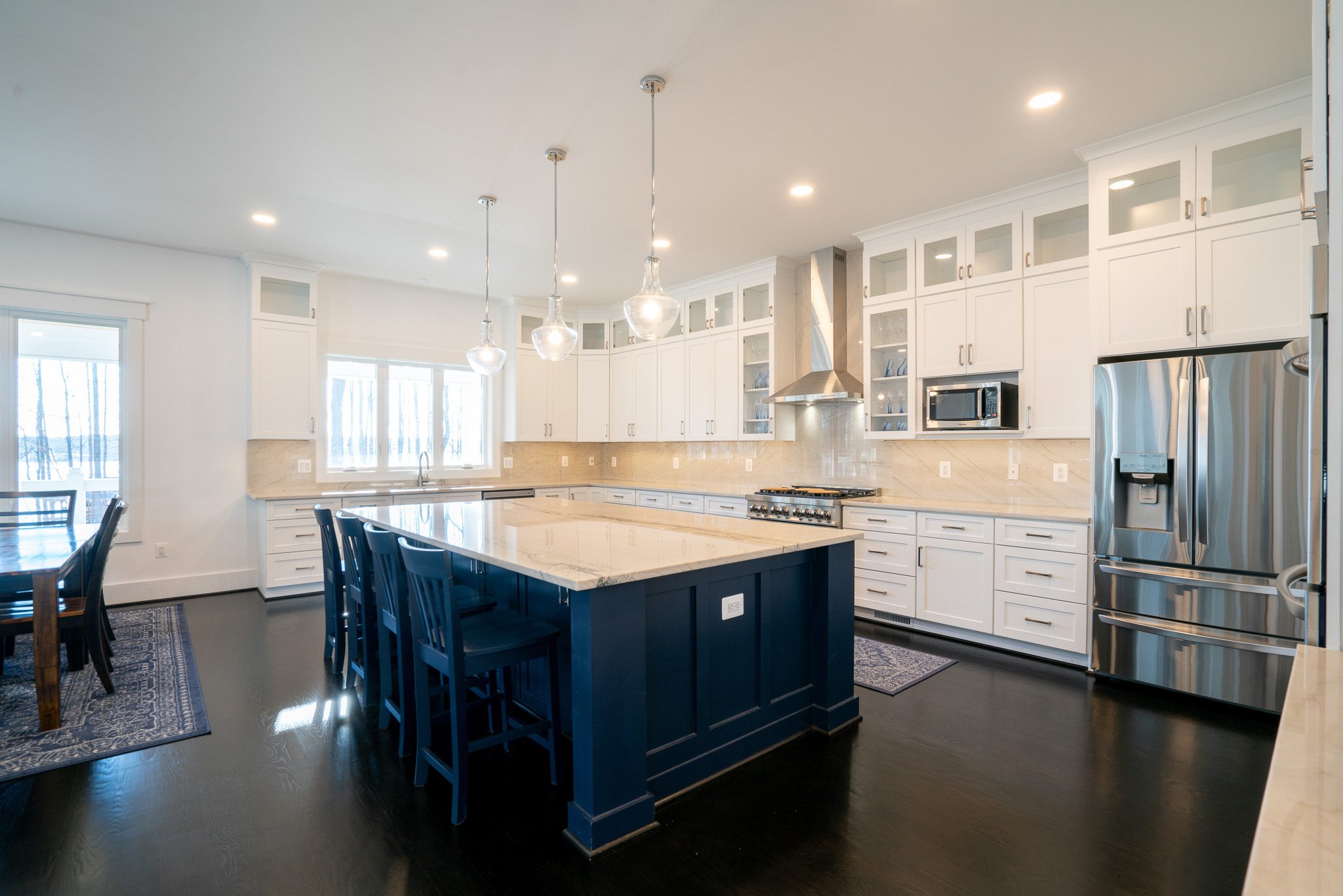 Modern kitchen with white cabinets, stainless steel appliances, a large island with a beige countertop, and dark hardwood floors. Pendant lights hang above the island.