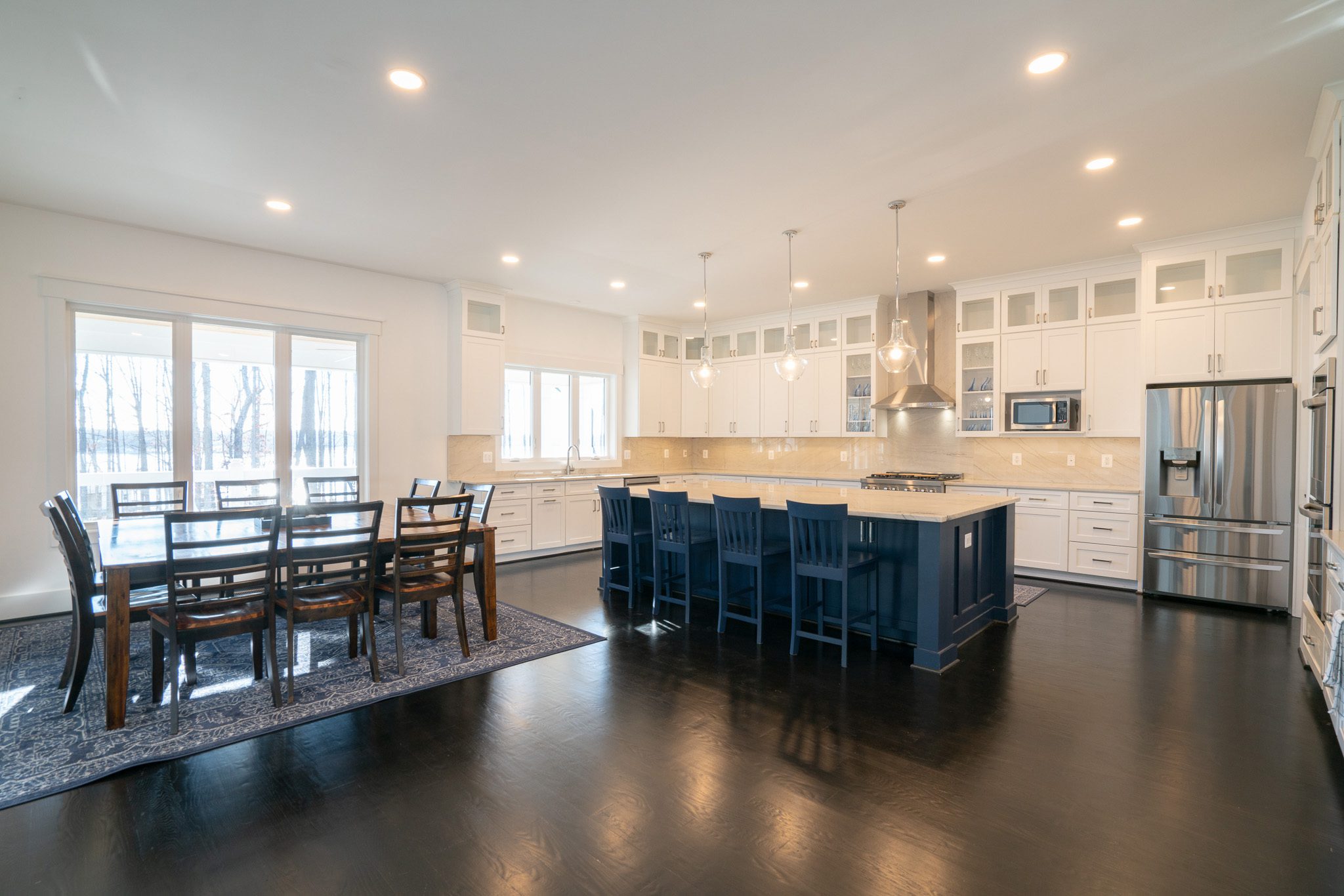Spacious kitchen with dark wood floors, large island with blue base, white cabinets, stainless steel appliances, pendant lights, and a dining table with chairs on a rug.