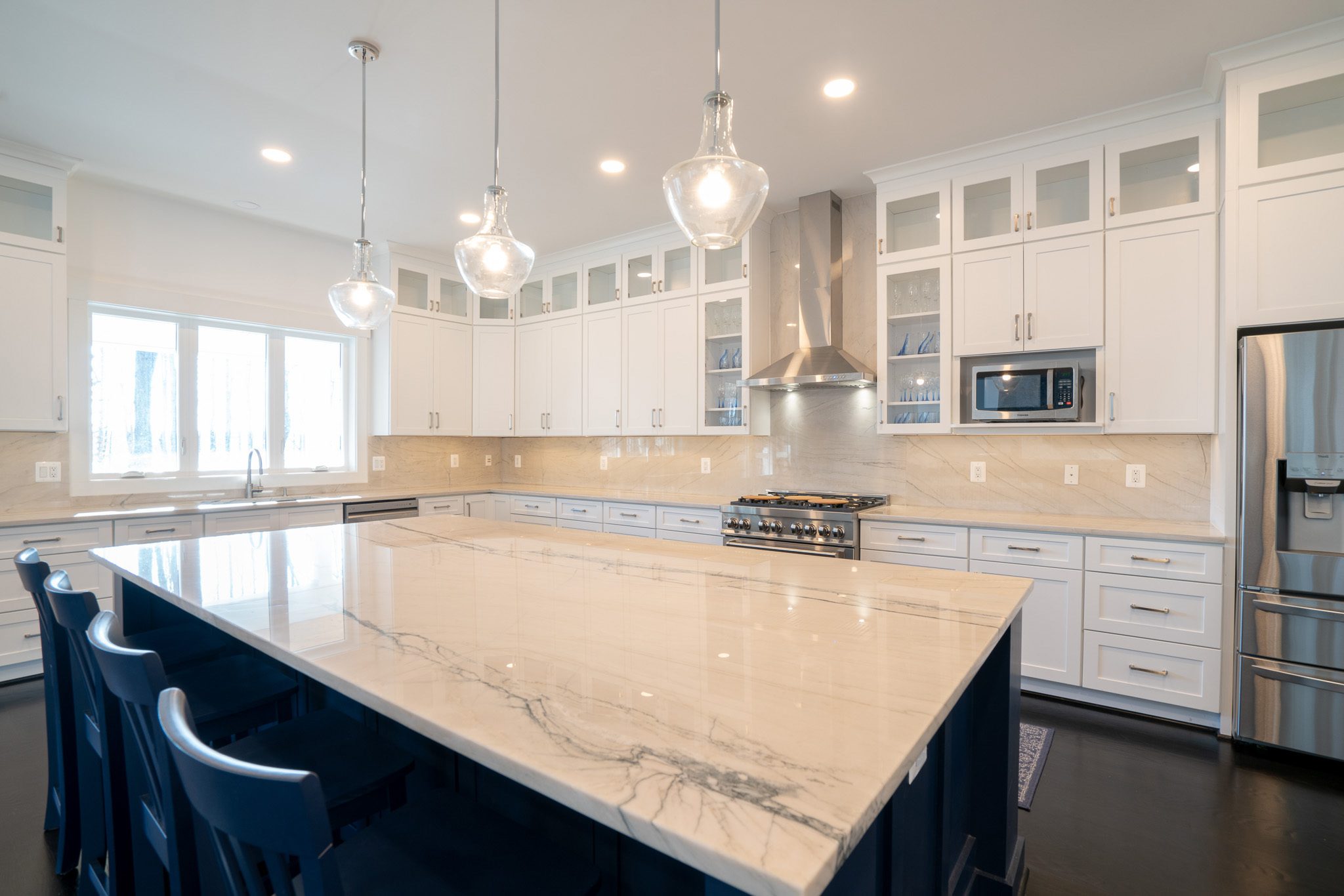 Modern kitchen with a large marble island, dark blue chairs, white cabinets, stainless steel appliances, and pendant lights.