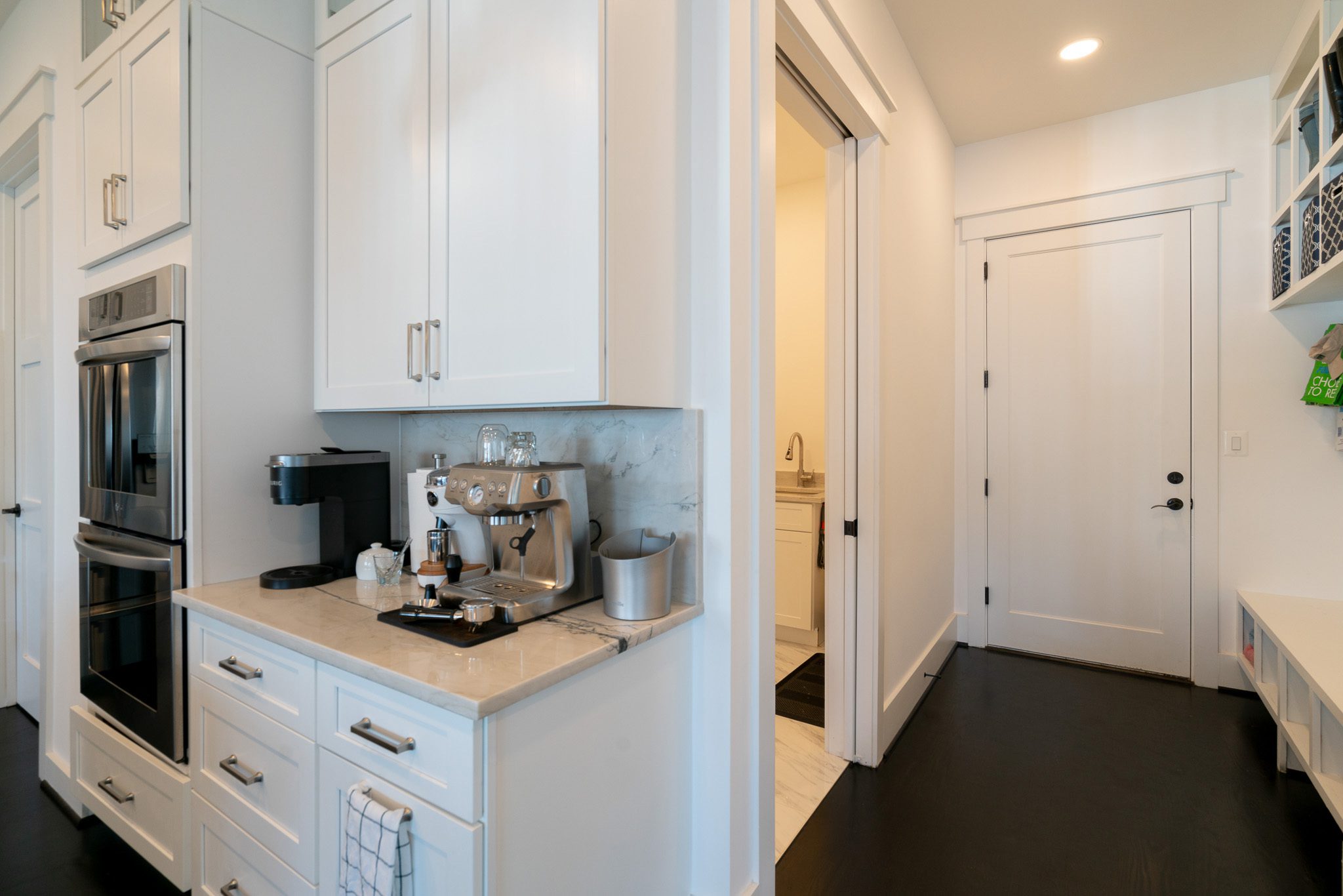 Modern kitchen with white cabinets, a double oven, a coffee maker, and an espresso machine on the counter. Nearby, a hallway leads to a door.