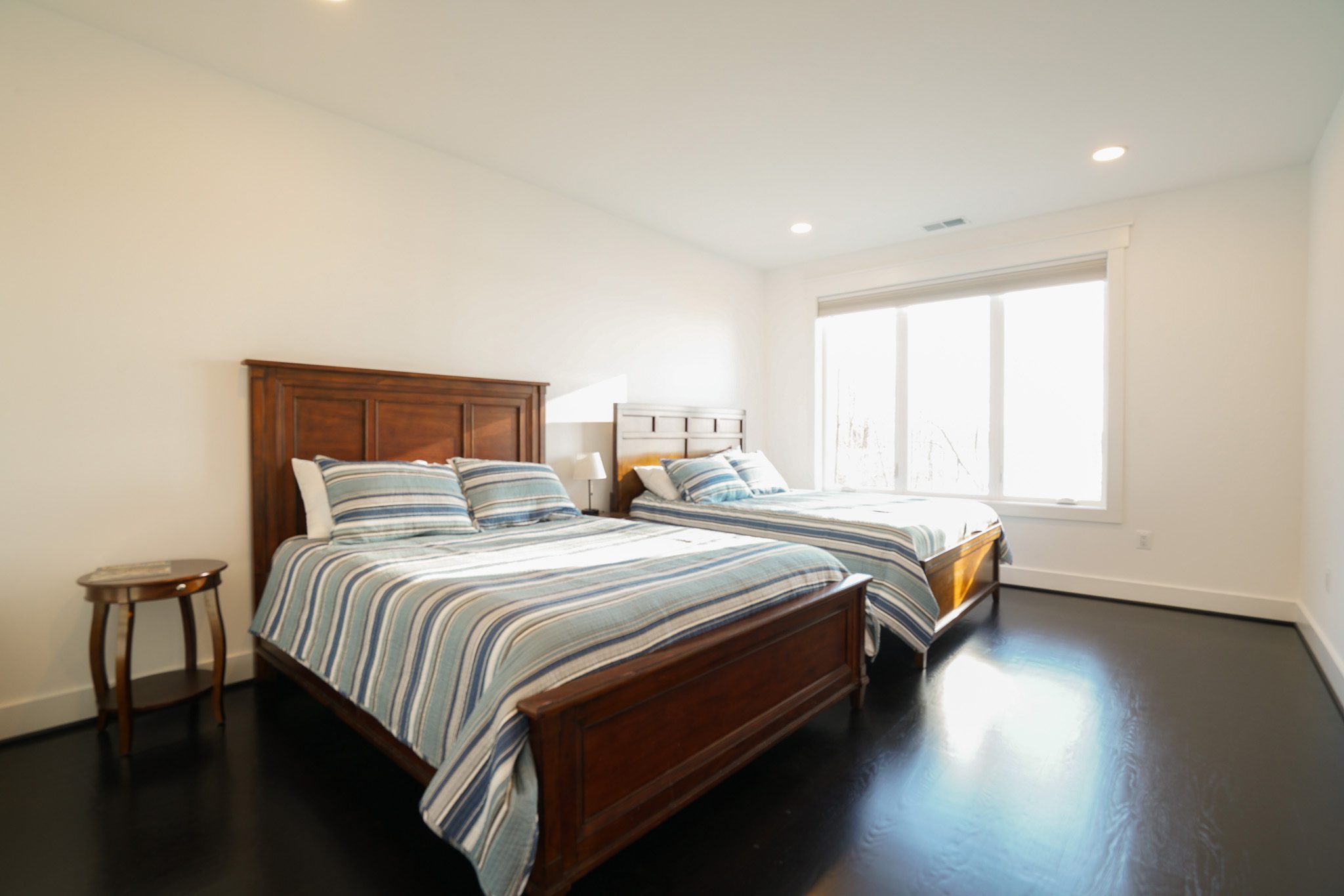 A minimalist bedroom features two wooden beds with striped bedding, a small round side table, and a large window letting in natural light. Walls and ceiling are white, and the floor is dark.