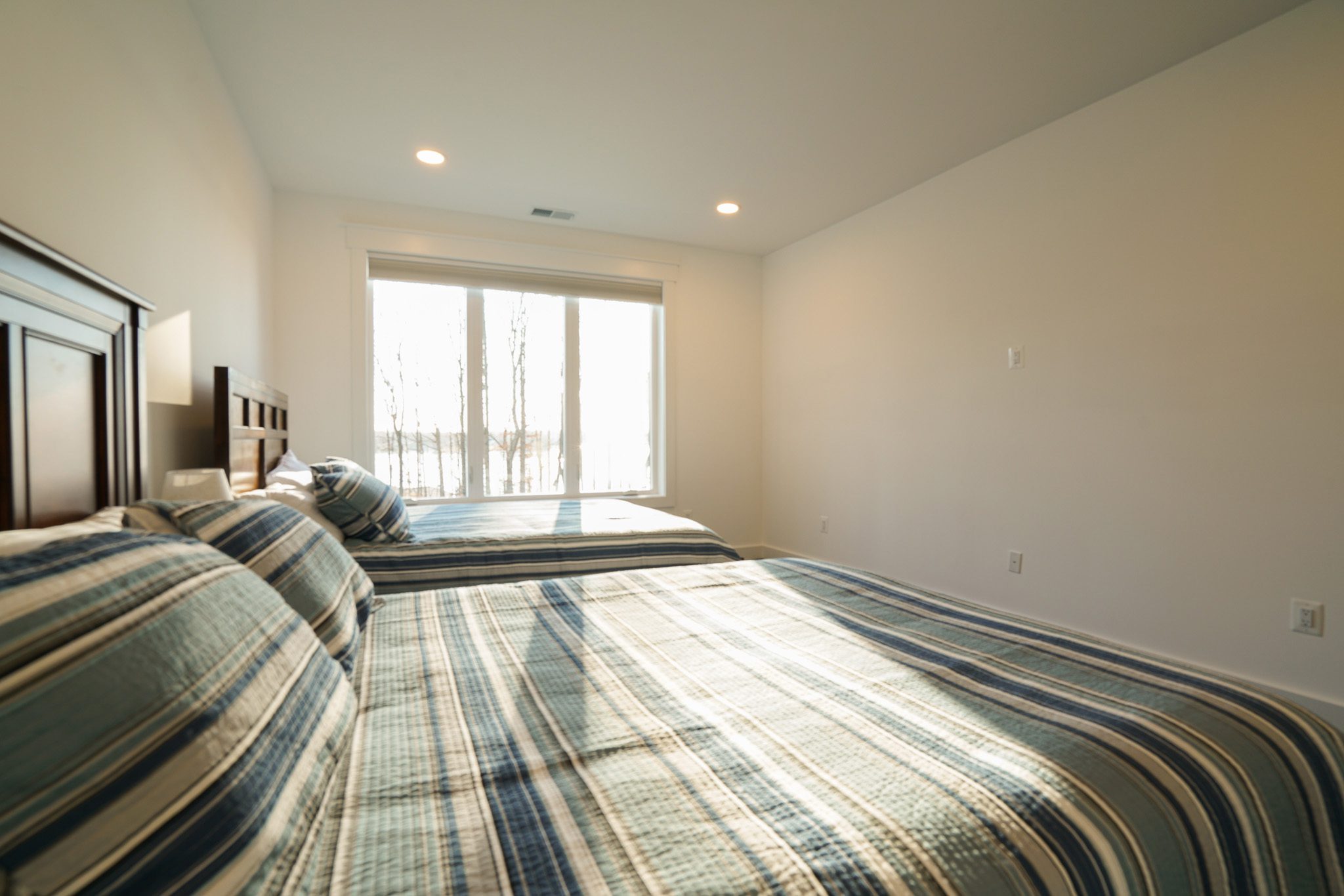 Two neatly made beds with striped bedding in a bright, minimalistic room featuring a large window and wooden headboard.