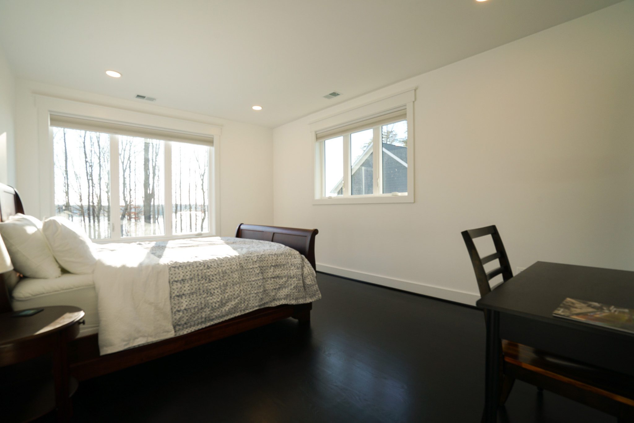 A bright bedroom with a large window, a bed with white and gray bedding, a small side table, and a dark wooden desk with a chair.