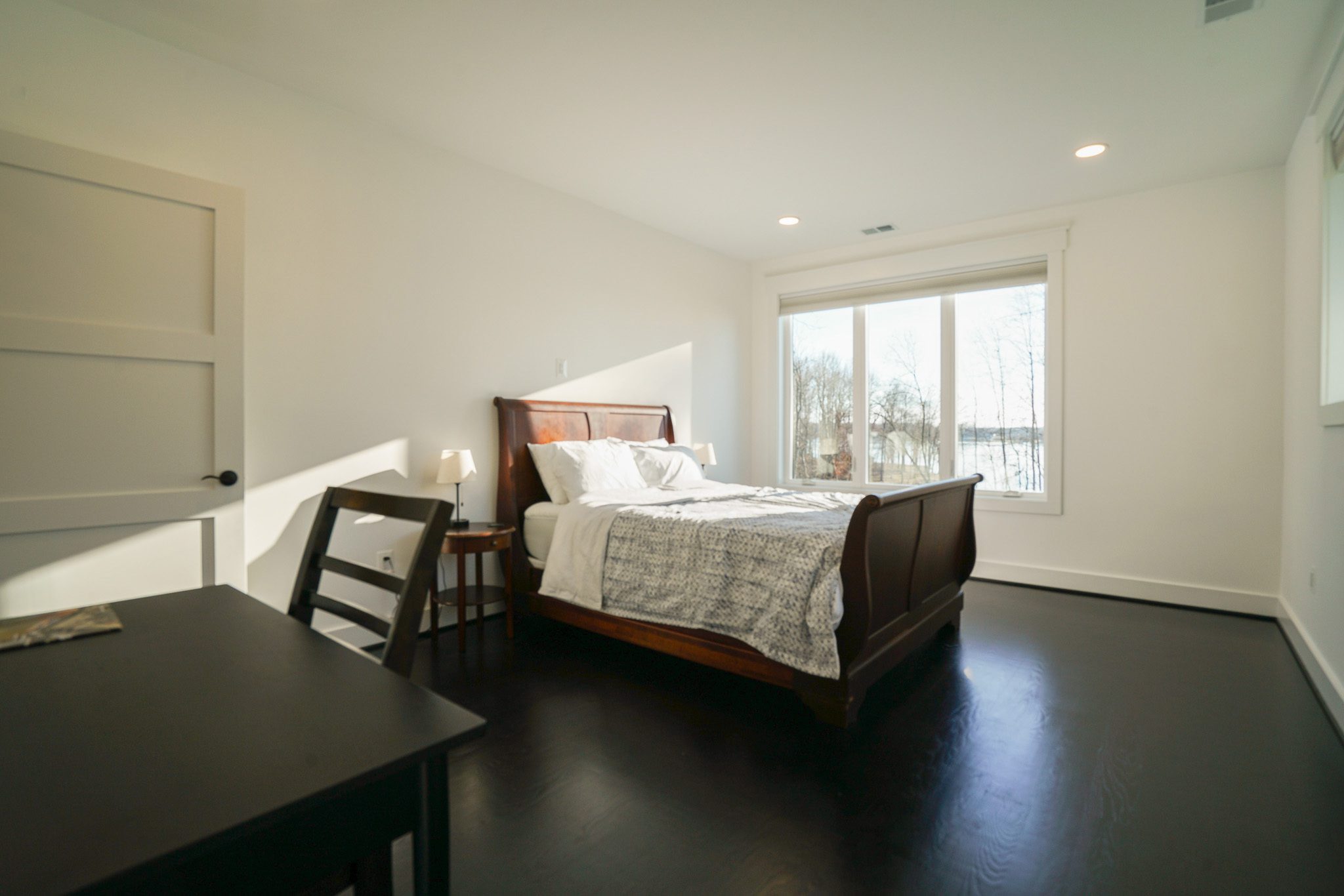 A minimalistic bedroom with a wooden bed, white bedding, a bedside lamp, and a small desk and chair. Large window showing a view of trees outside.