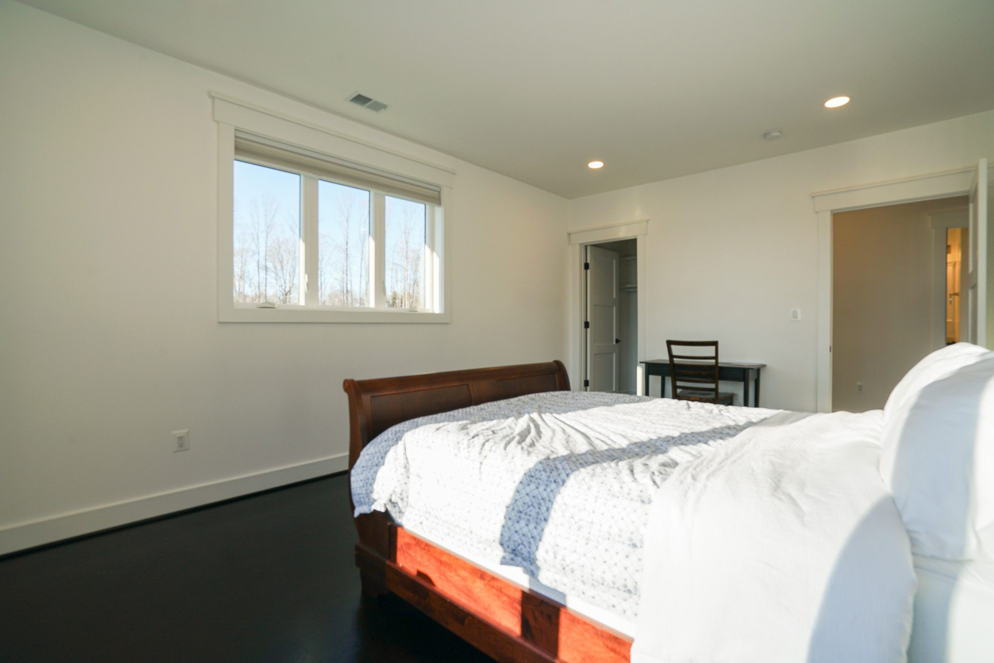 Bright bedroom with a sleigh bed, white bedding, a small desk, and a chair. Window with a view of trees, light walls, and dark flooring.