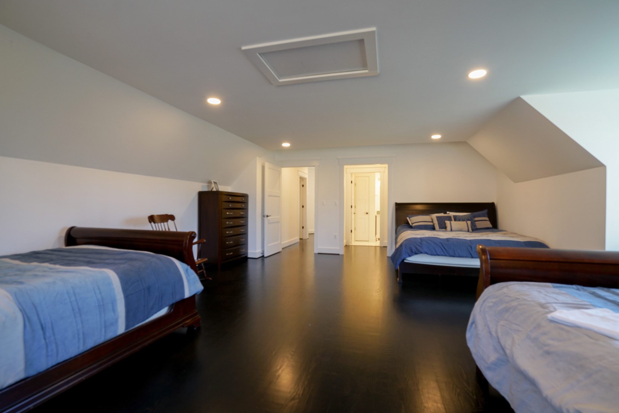 Bedroom with three beds, dark wood flooring, and light-colored walls. Dresser and rocking chair on the left, and recessed lighting in the ceiling.