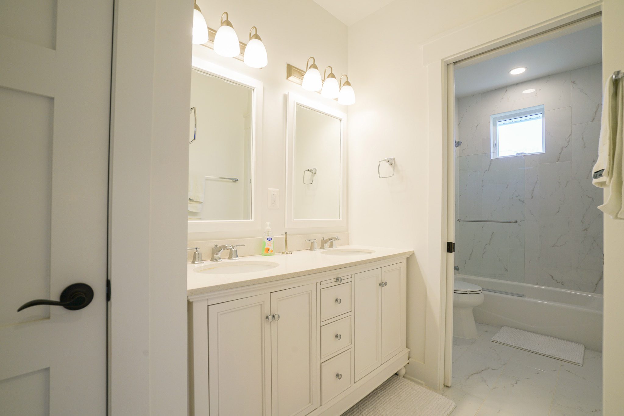 Bright bathroom with white double sink vanity, two mirrors, and overhead lighting. Shower area with marble tiles visible through open door. White walls and fixtures throughout.