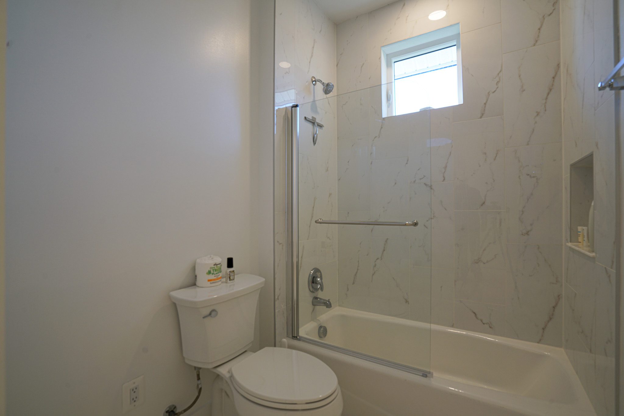 Bathroom featuring a white toilet, bathtub with a glass door, and beige marble-patterned tiles on the wall. A small window is above the bathtub.