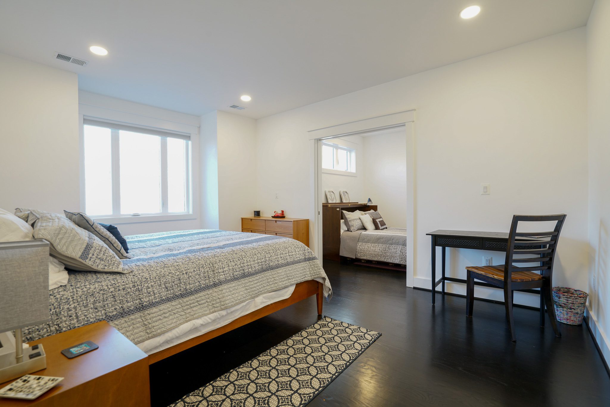 Modern bedroom with a large bed, dark wood flooring, a small desk with a chair, and a visible second bedroom through an open sliding door. Walls are painted white, and lighting is recessed.