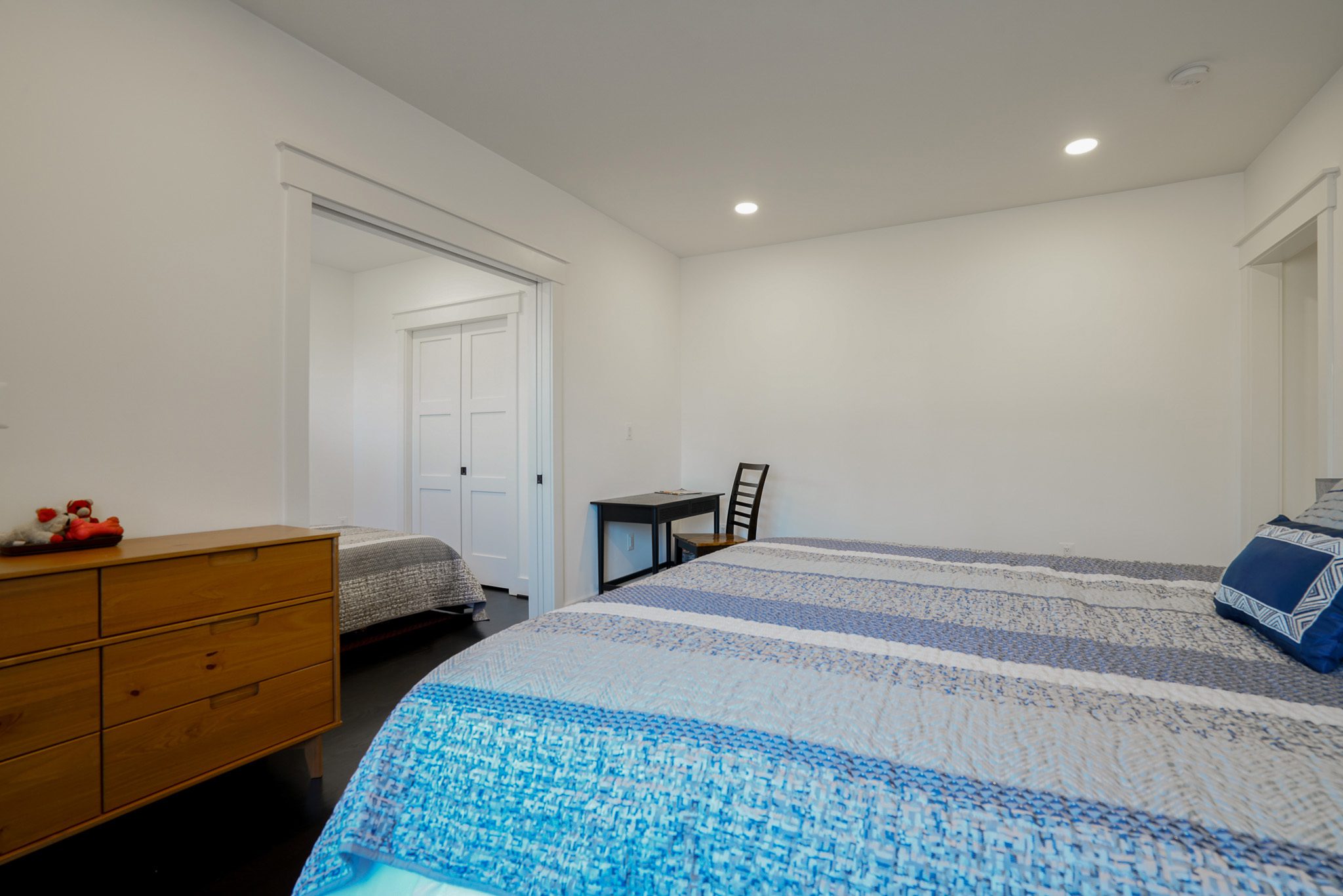 Bedroom with a bed featuring blue and white striped bedding, a wooden dresser, a small desk with a chair, and an adjacent room visible through a doorway.