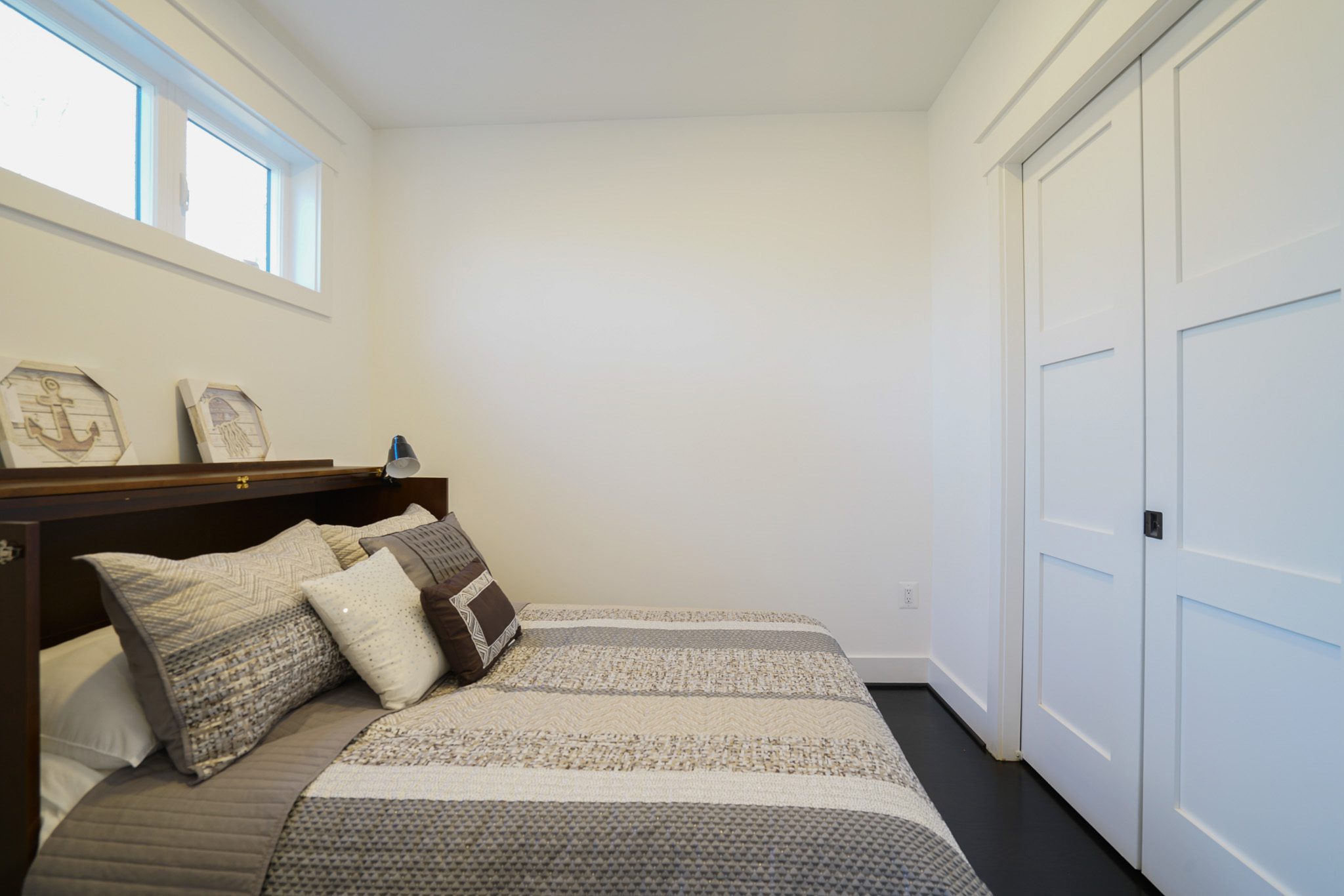 A small bedroom with a neatly made bed adorned with gray and white bedding. There are decorative items on a shelf above the bed, and a sliding double door closet is on the right wall.