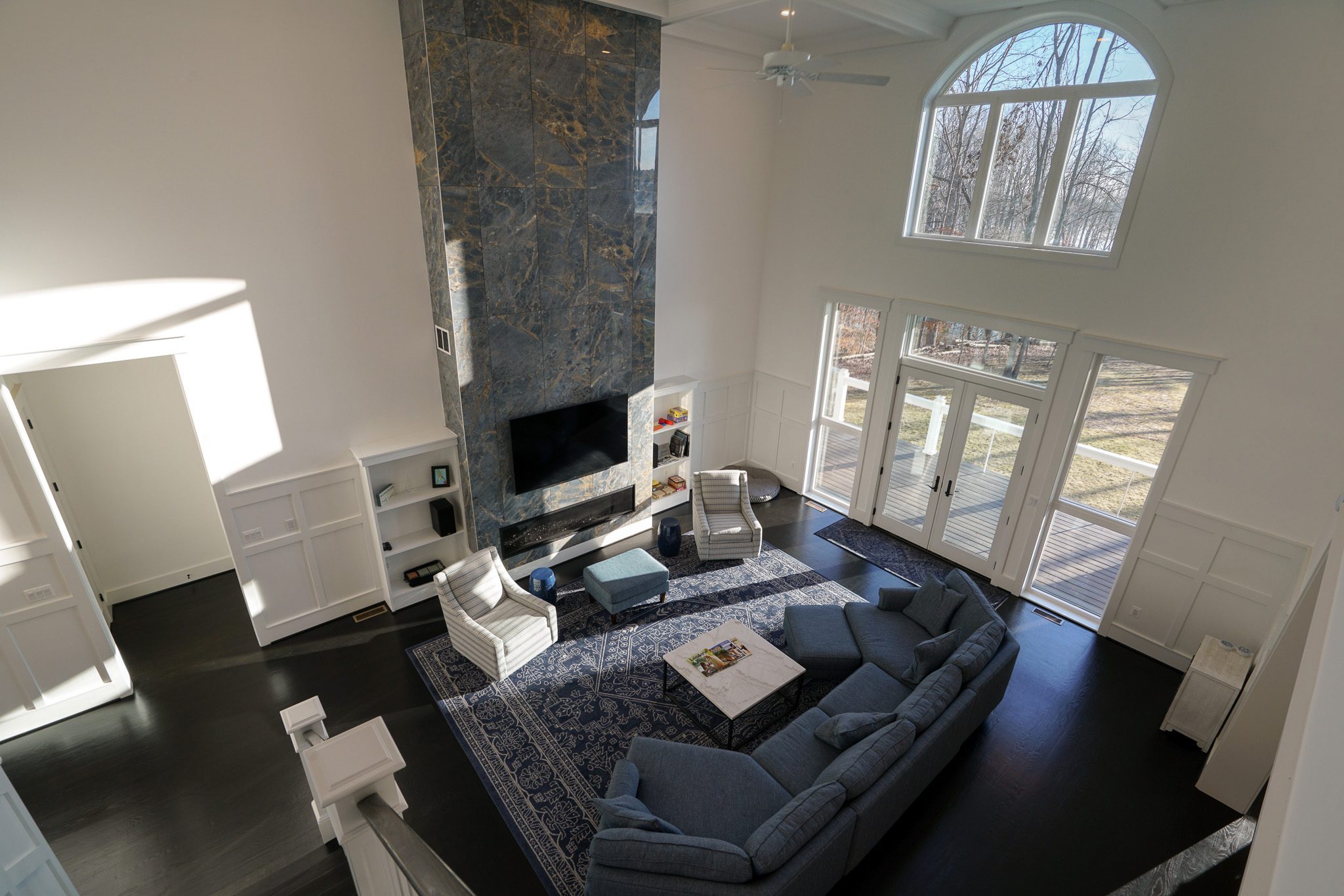 A spacious living room with a high ceiling, large gray sectional sofa, two armchairs, a TV on a stone feature wall, and floor-to-ceiling windows overlooking a wooden deck and trees.
