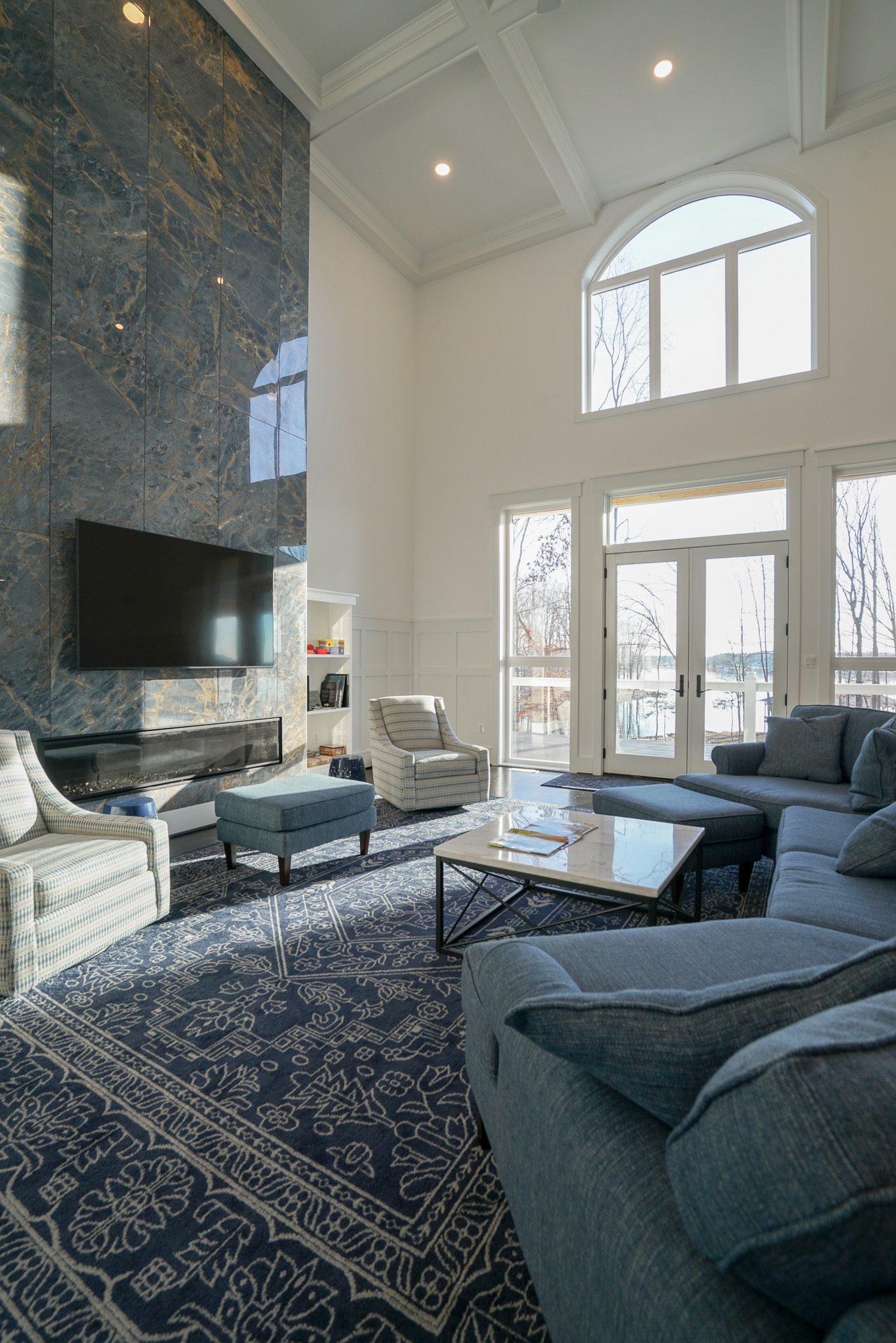 Spacious living room with high ceilings, large windows, blue-gray furniture, a patterned rug, and a wall-mounted TV above a tall stone fireplace.