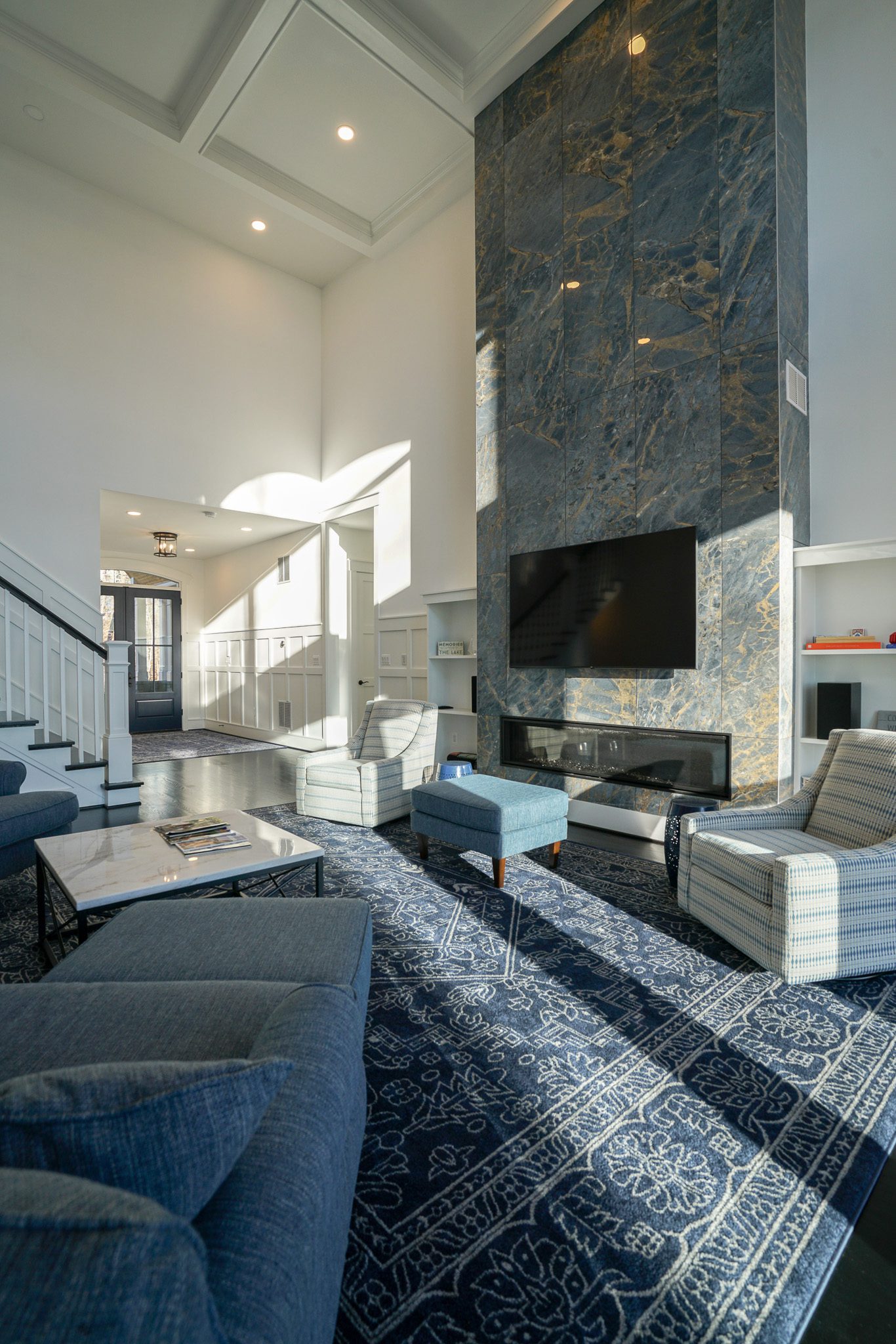 Spacious living room with tall ceiling, large stone fireplace, mounted TV, blue sofas, chairs, and patterned rug. Staircase leads to upper floors, and sunlight streams through windows.