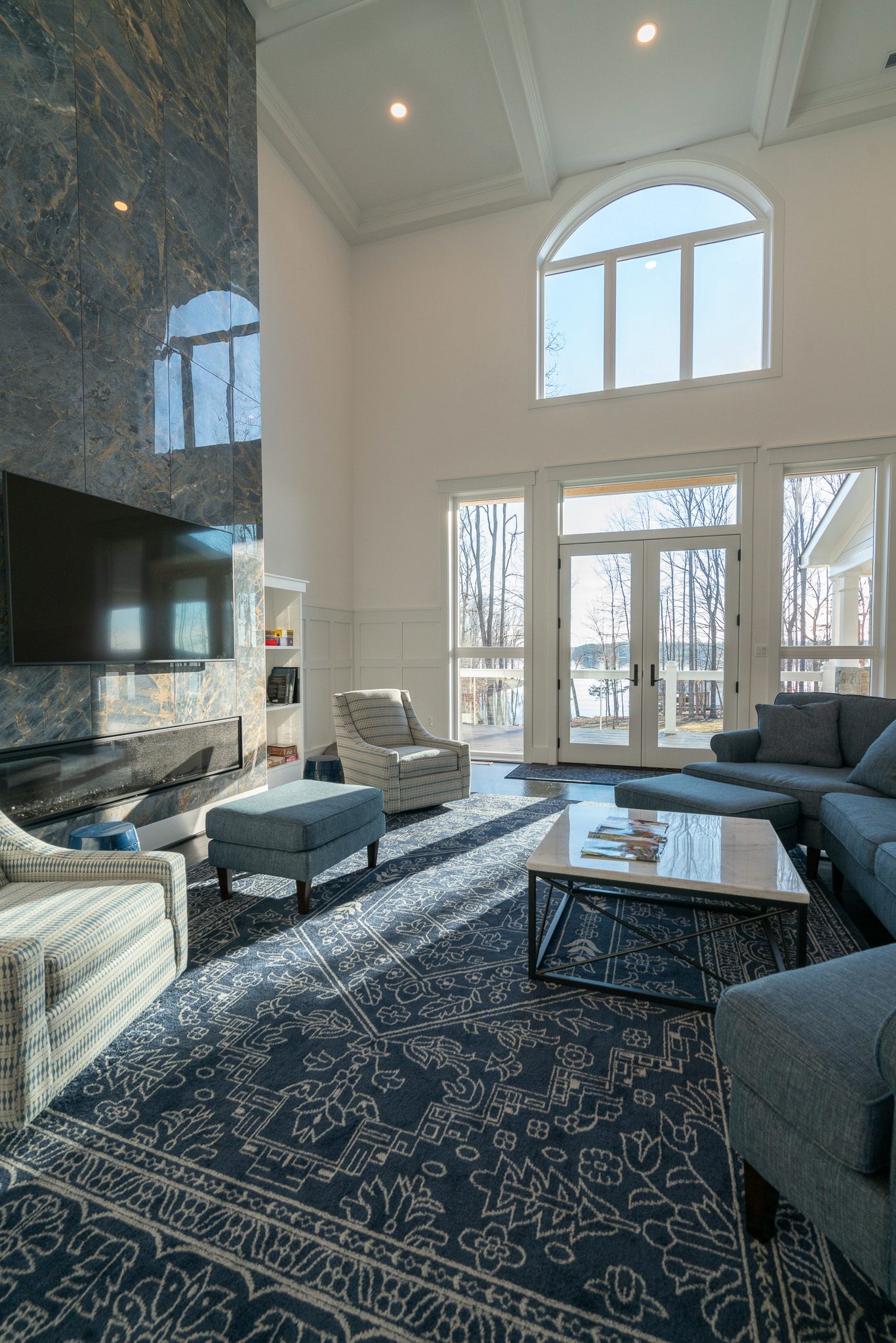 A spacious living room with high ceilings, featuring blue patterned rugs, a large wall-mounted TV, a marble fireplace, and several modern chairs and sofas under natural light from tall windows.