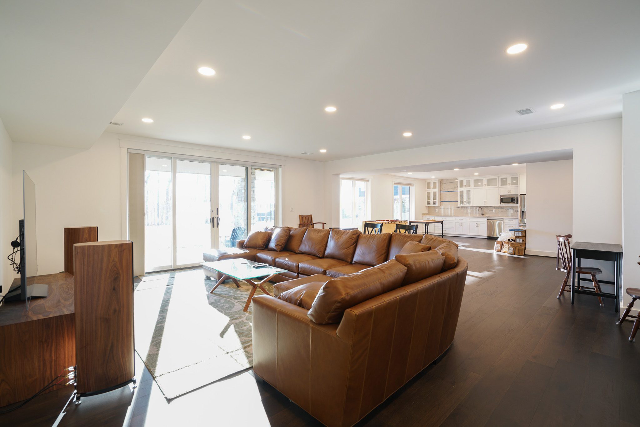 Open living room with brown leather sectional sofa, wooden coffee table, and large windows. Modern kitchen with white cabinets in the background. Dark hardwood flooring throughout.