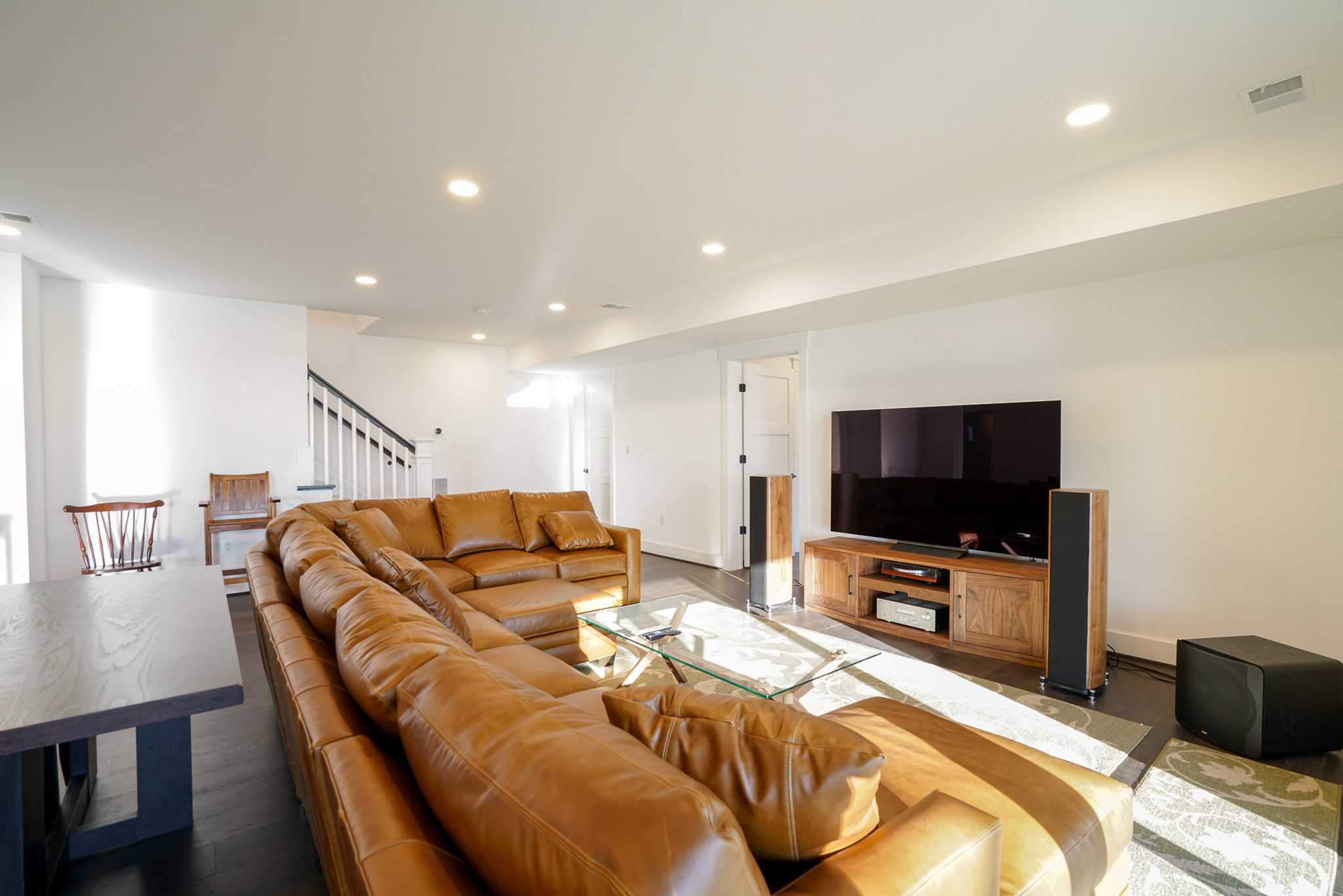 Spacious living room with a large brown leather sectional sofa, glass coffee table, flat-screen TV on a wooden stand, and a staircase in the background. Bright, natural lighting fills the room.