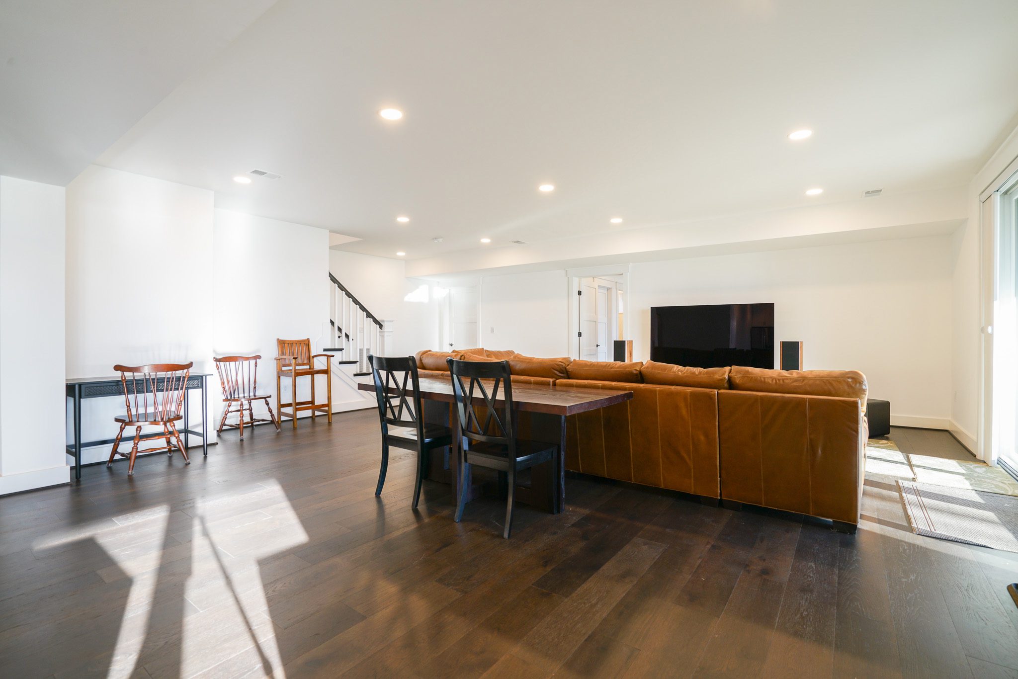 Spacious living room with a brown leather sectional sofa, dining table with chairs, flat-screen TV, and wooden flooring, illuminated by recessed lighting and natural light from windows.