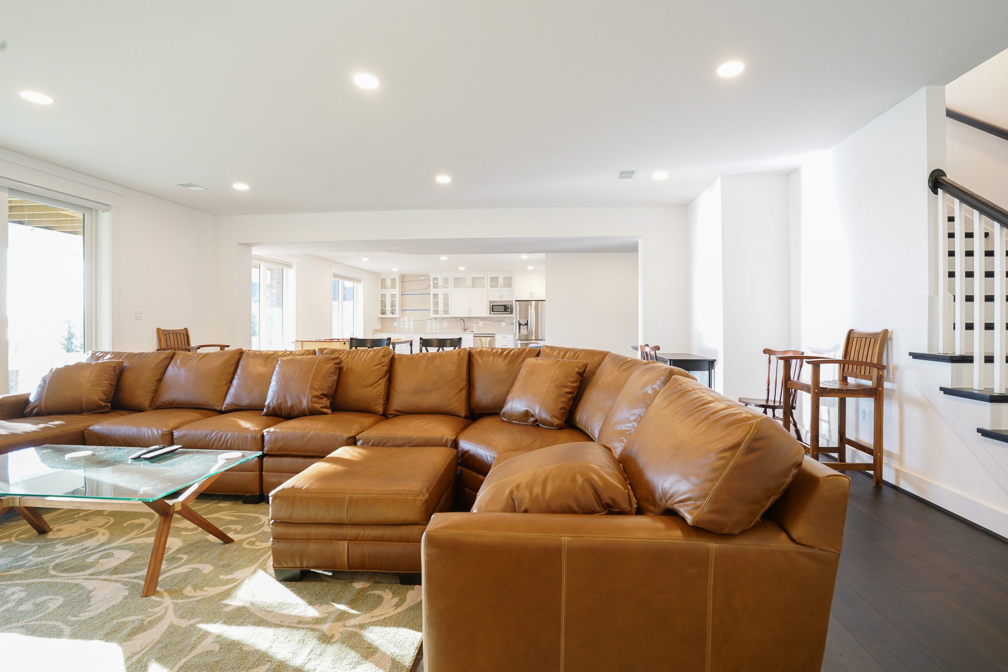 Living room with a large brown leather sectional sofa, glass coffee table, and open view of a bright kitchen in the background. Stairs and barstools are on the right.