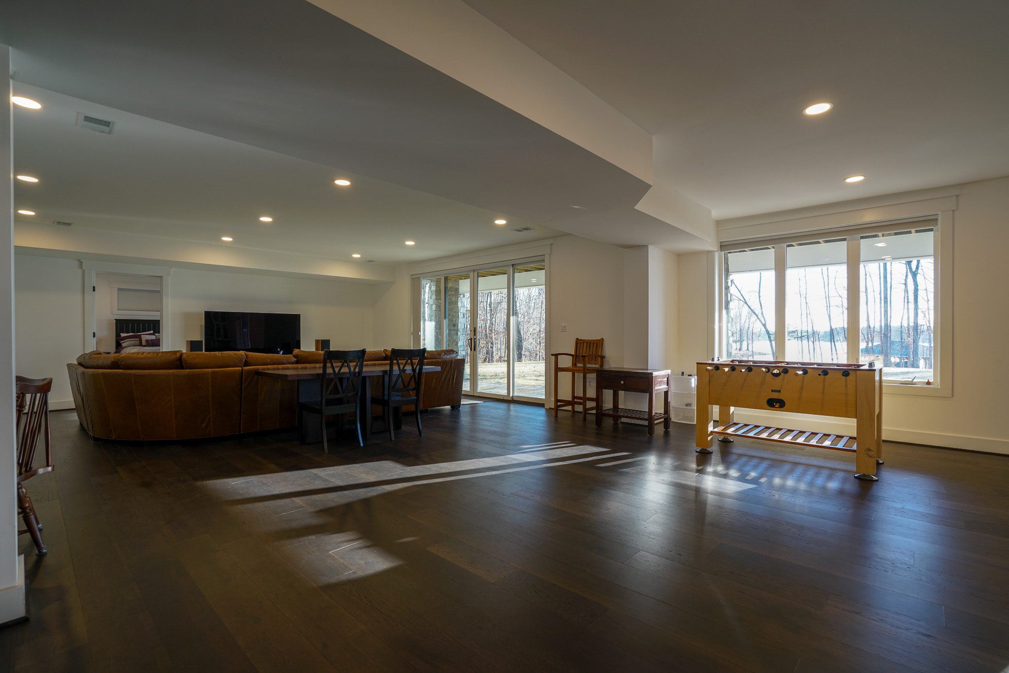 Spacious living room with dark wood flooring, a large brown sectional sofa, a TV, a round dining table with chairs, and a foosball table near large windows.